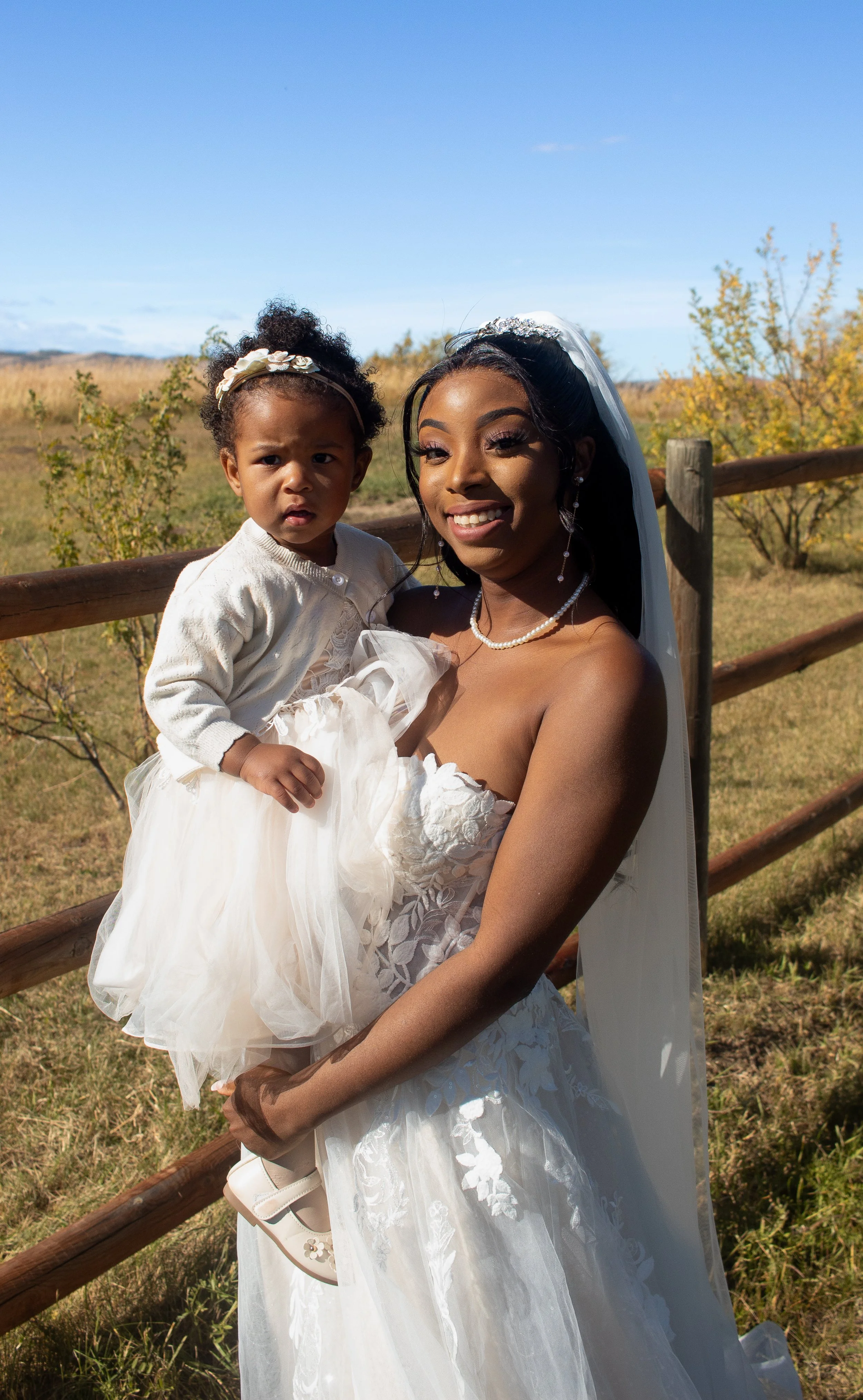 A woman dressed in a wedding gown holding a young girl in a cream-colored dress outdoors near a wooden fence, with trees and a clear blue sky in the background.