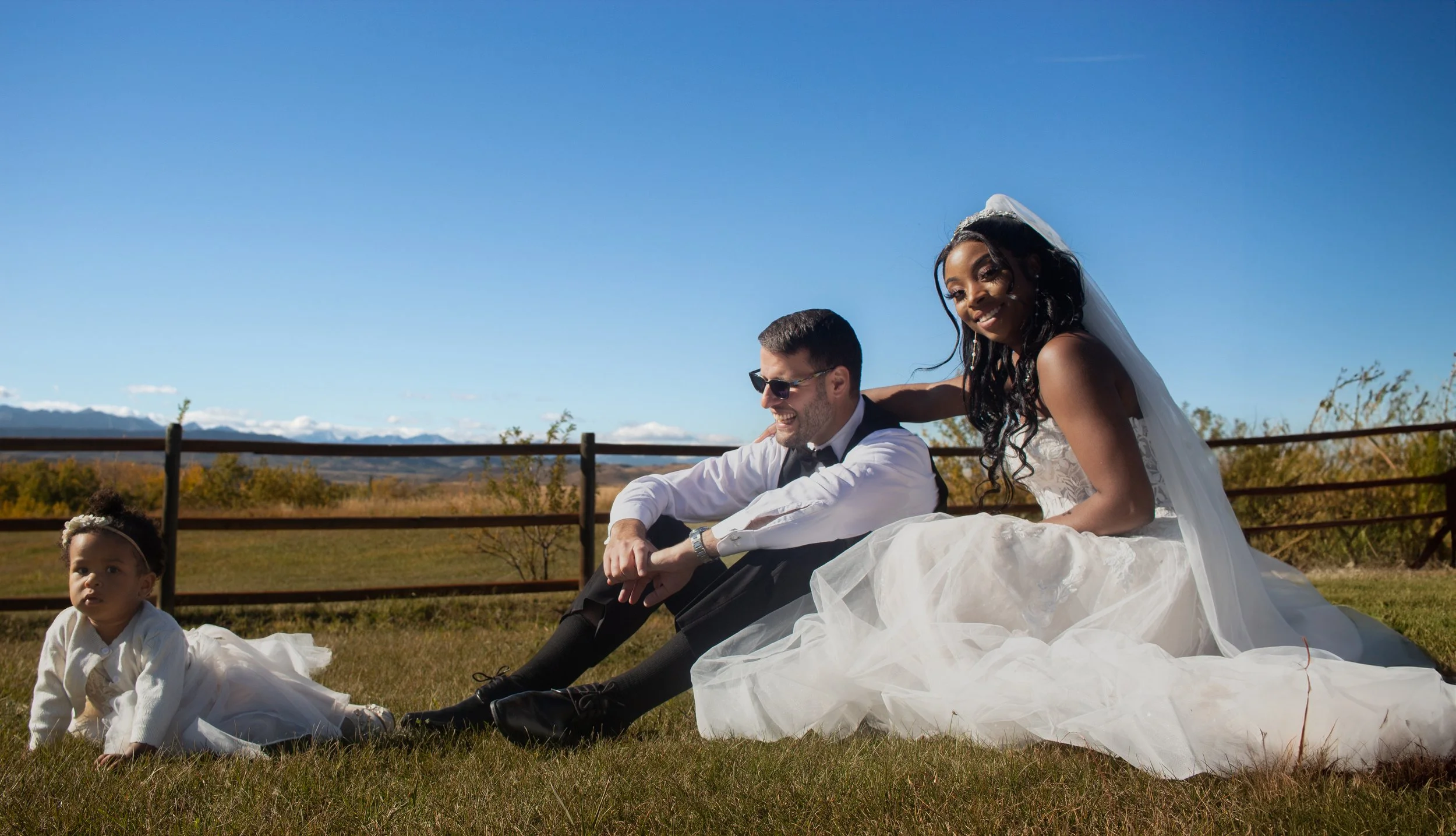 A bride and groom sitting outdoors on grass during a sunny day, with a blue sky and distant mountains in the background. The bride is wearing a white wedding dress and veil, smiling, and leaning towards the groom. The groom is wearing sunglasses, a t