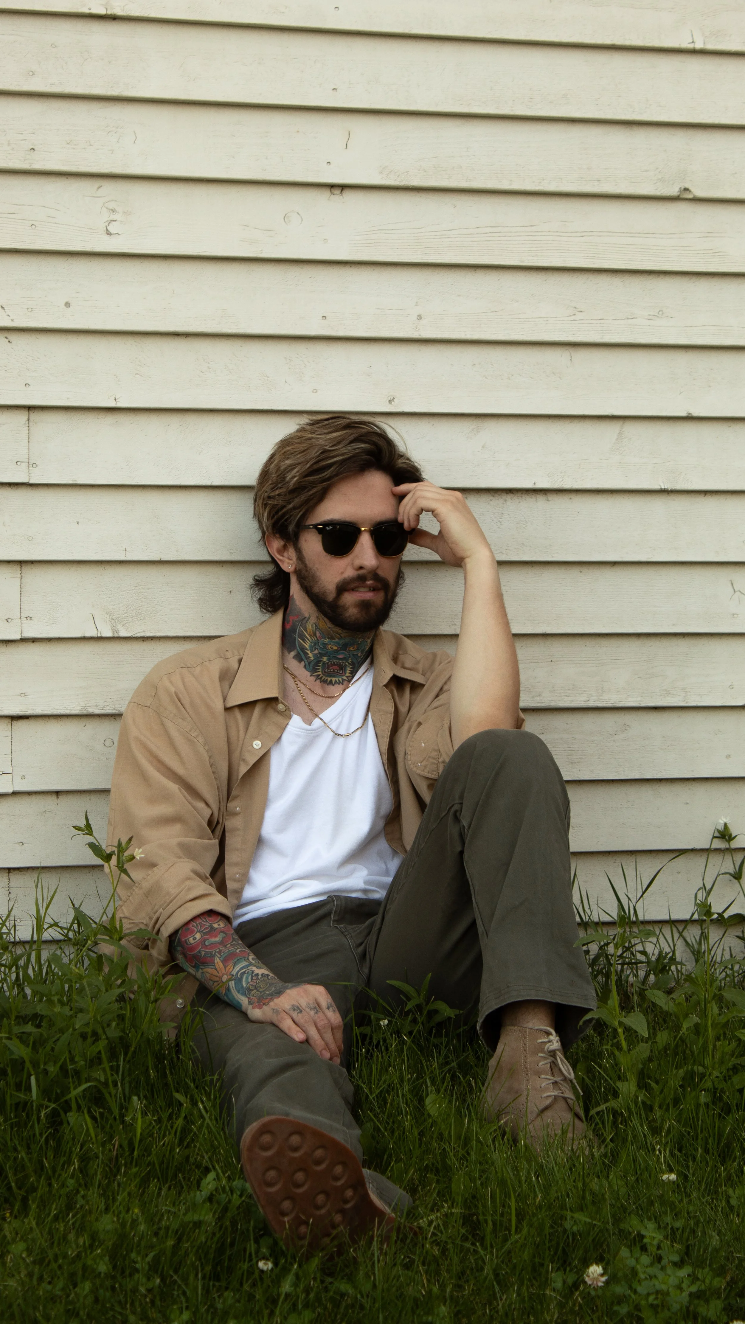 A man with brown hair, beard, and tattoos on his neck and arms, wearing sunglasses, a beige open shirt over a white t-shirt, dark pants, and tan boots, sitting on grass in front of a white wooden wall.