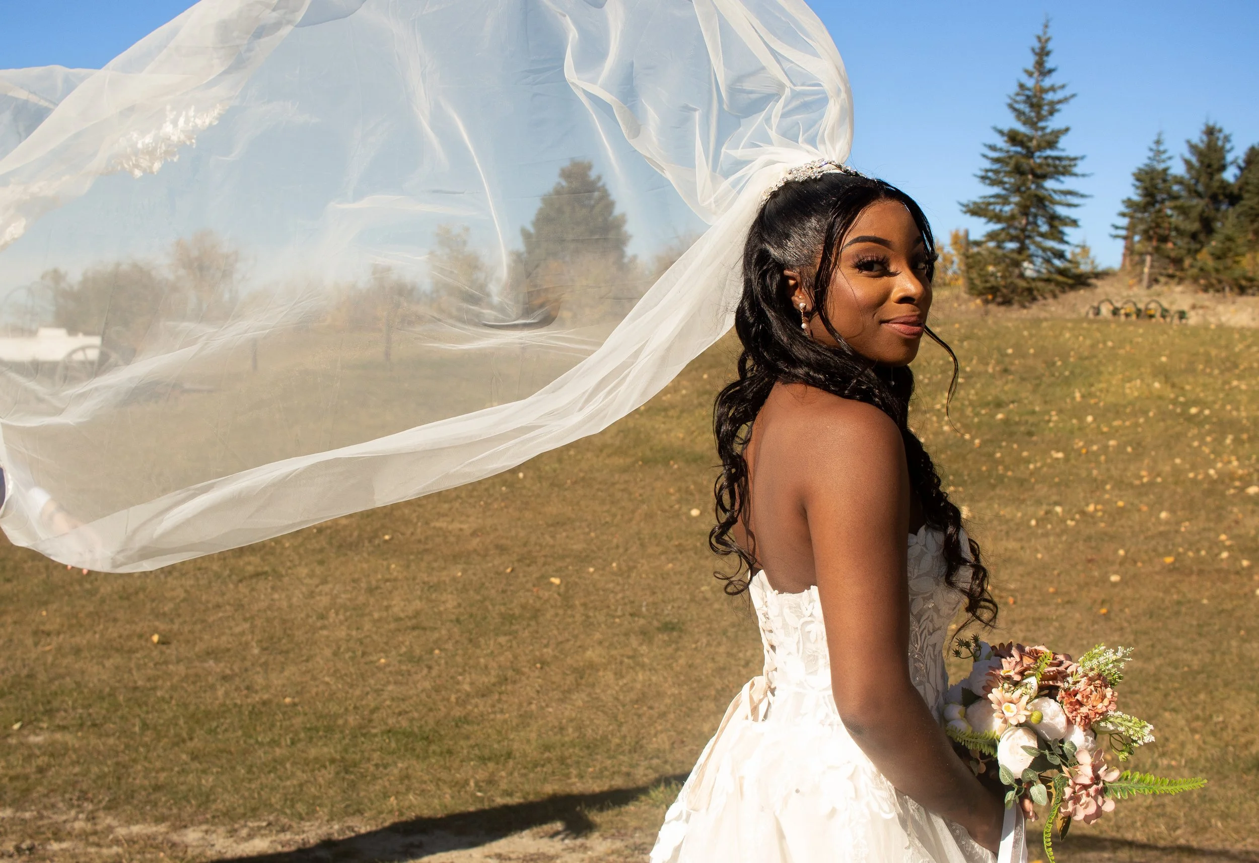A woman wearing a wedding dress and veil, holding a bouquet of flowers outdoors with trees and a clear blue sky in the background.