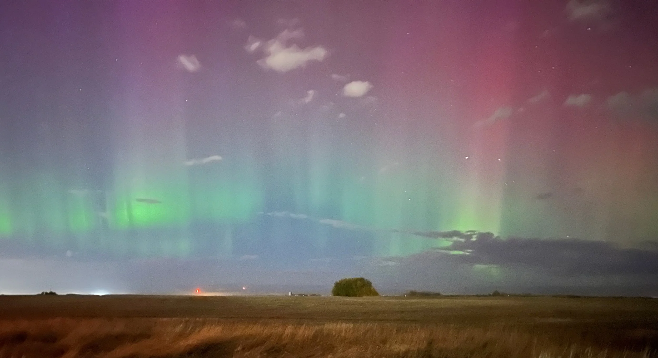Nighttime landscape showing the Northern Lights with green and purple hues across the sky, scattered clouds, and a flat horizon with sparse vegetation.