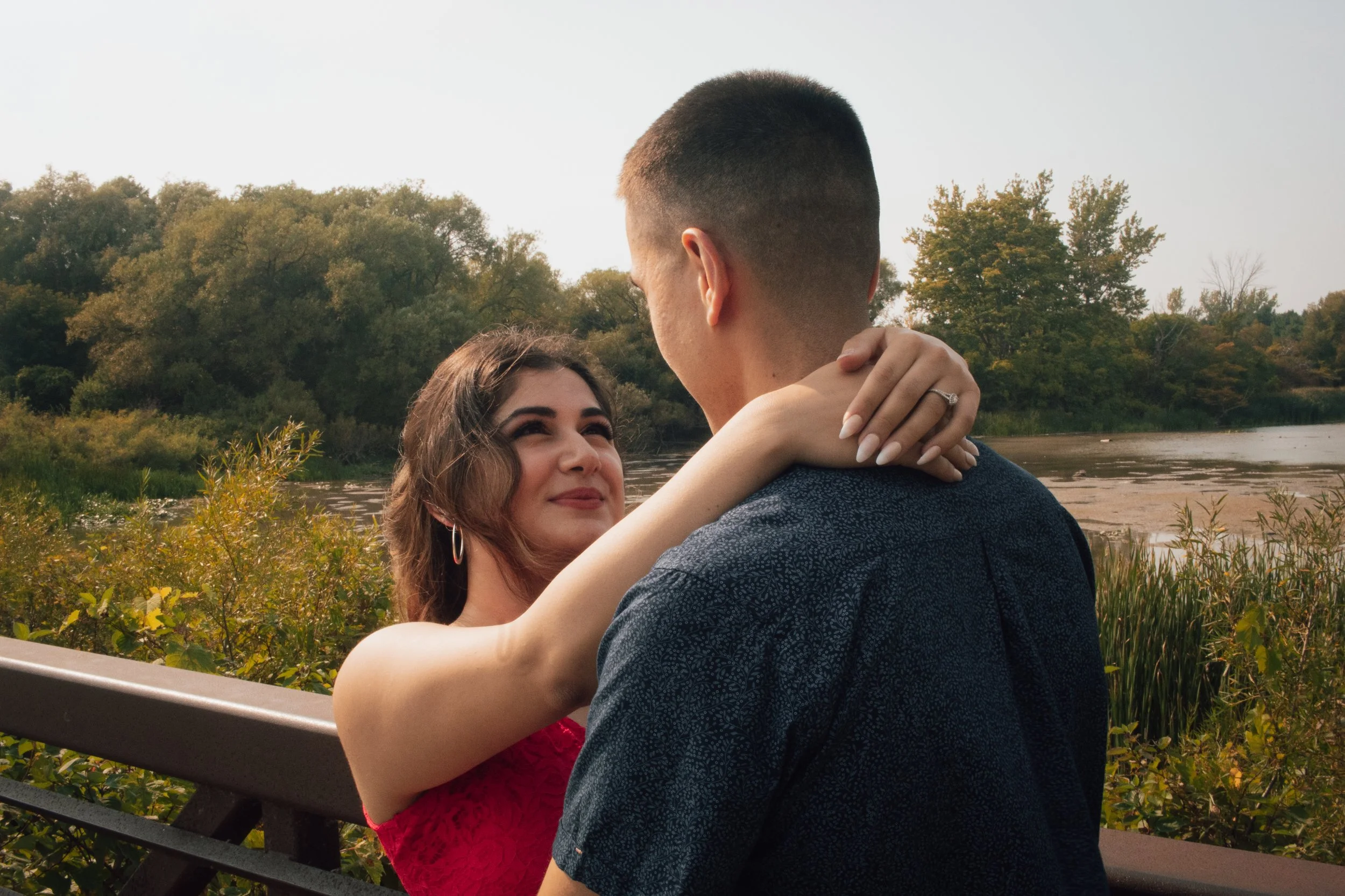 A man and woman embracing outdoors near a river with trees in the background during daytime.
