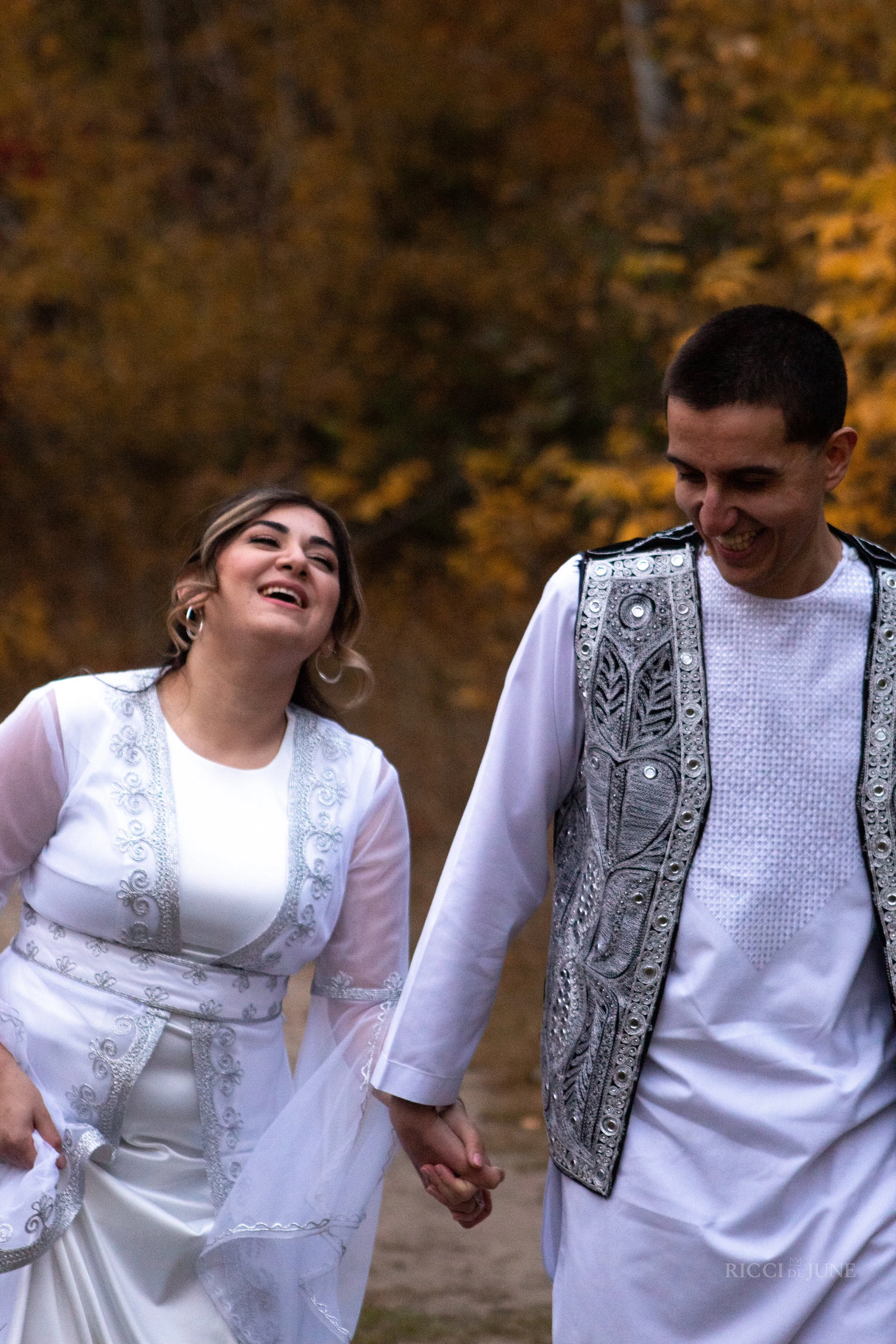 A couple holding hands, dressed in white traditional attire with intricate embroidery, walking outdoors during autumn, with trees displaying fall foliage in the background.