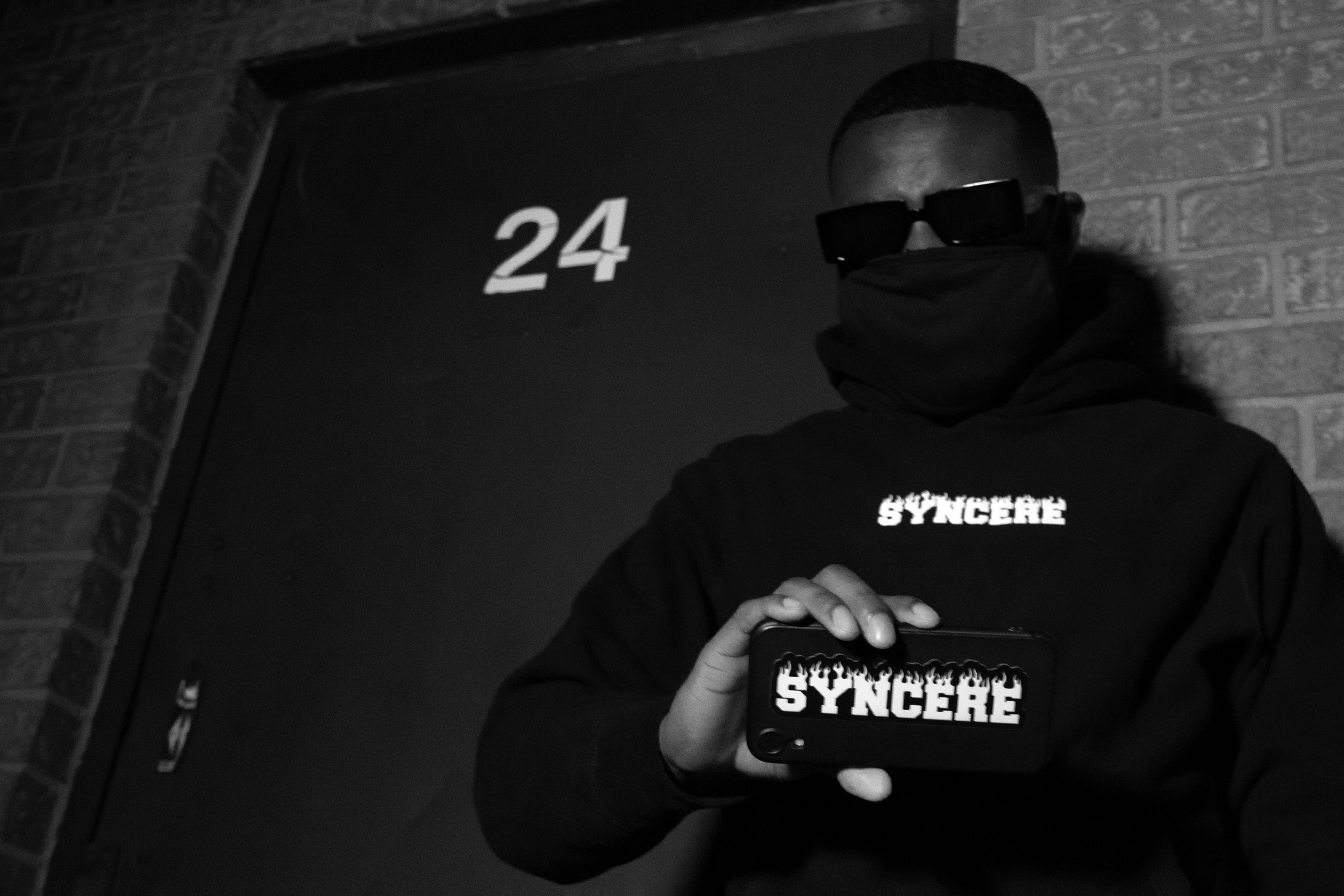 Person taking a mirror selfie wearing dark sunglasses and a hoodie with the word 'SYNCRERE' on it, standing near a brick wall and a door with the number '24'.