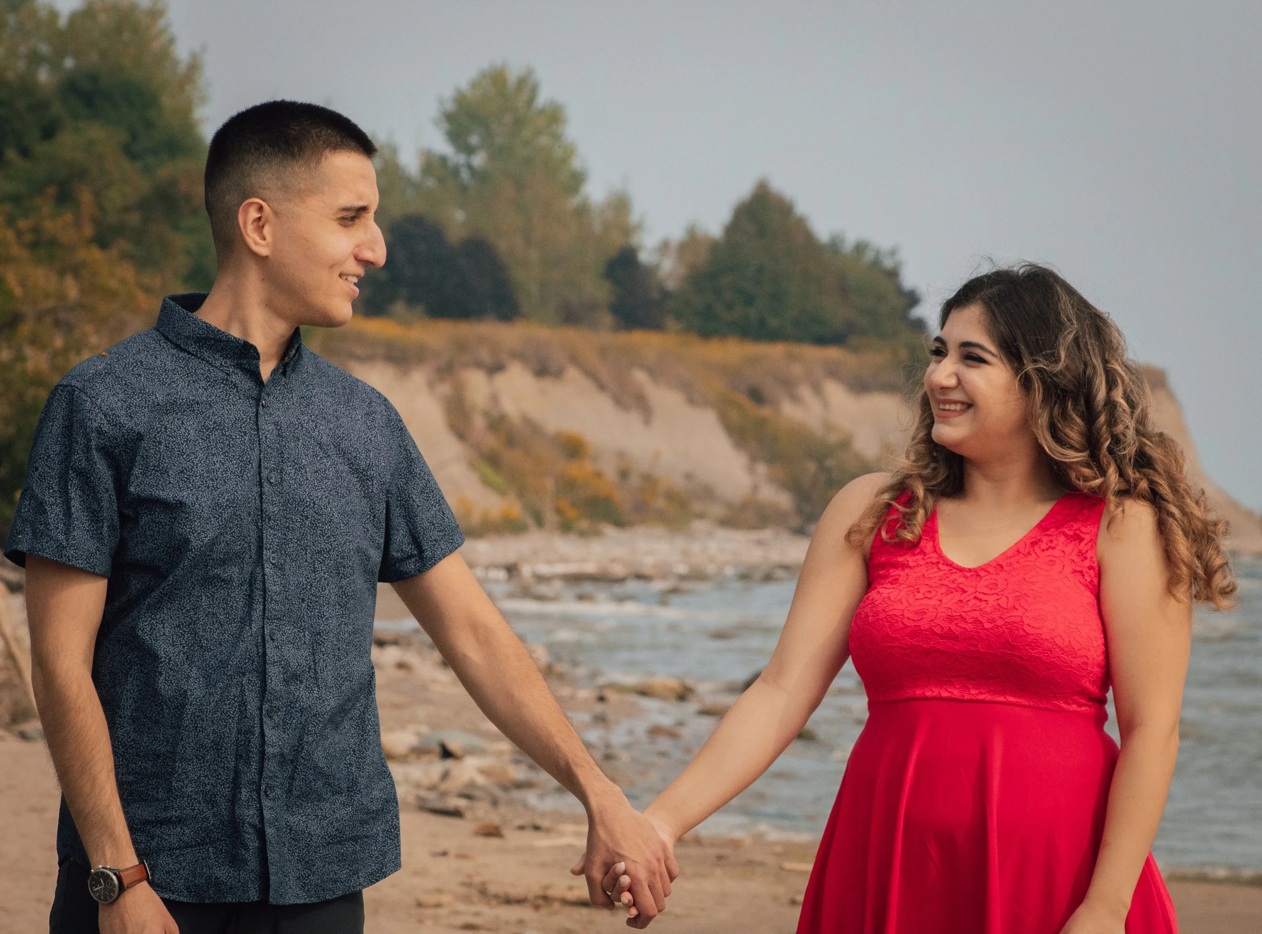 A young man and woman holding hands on the beach, smiling at each other with a shoreline and trees in the background.