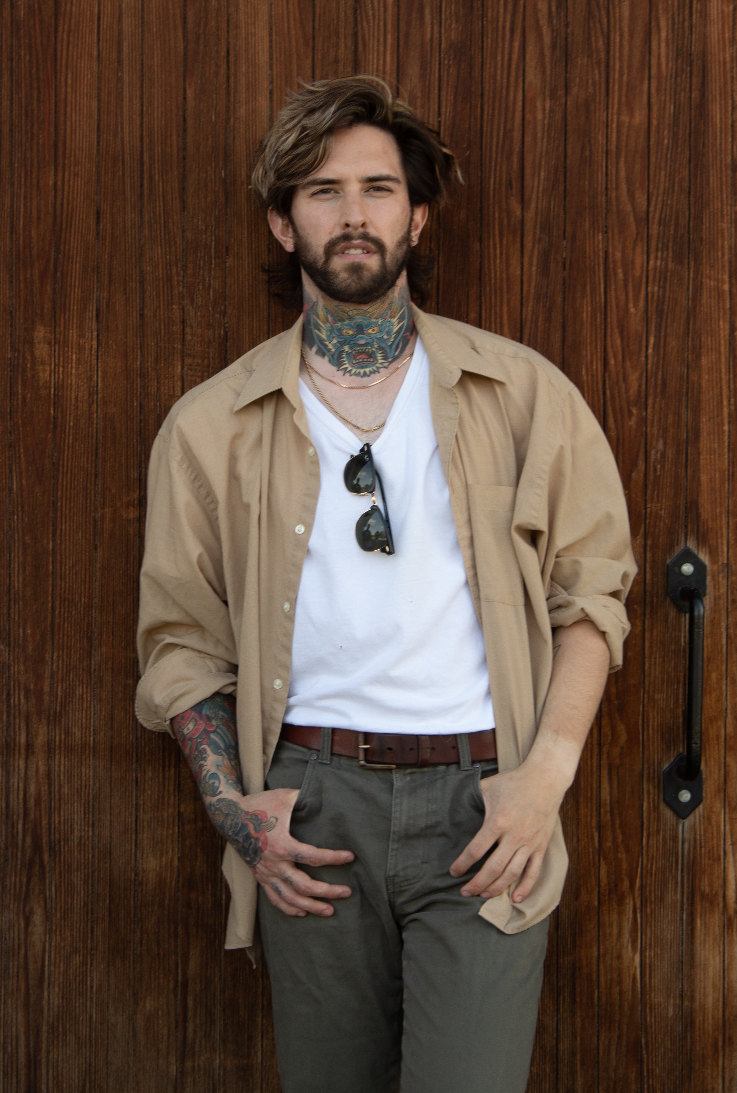 Man with tattoos, beard, and tousled hair leaning against a wooden wall, wearing a beige shirt over a white t-shirt, with sunglasses hanging from the shirt collar.