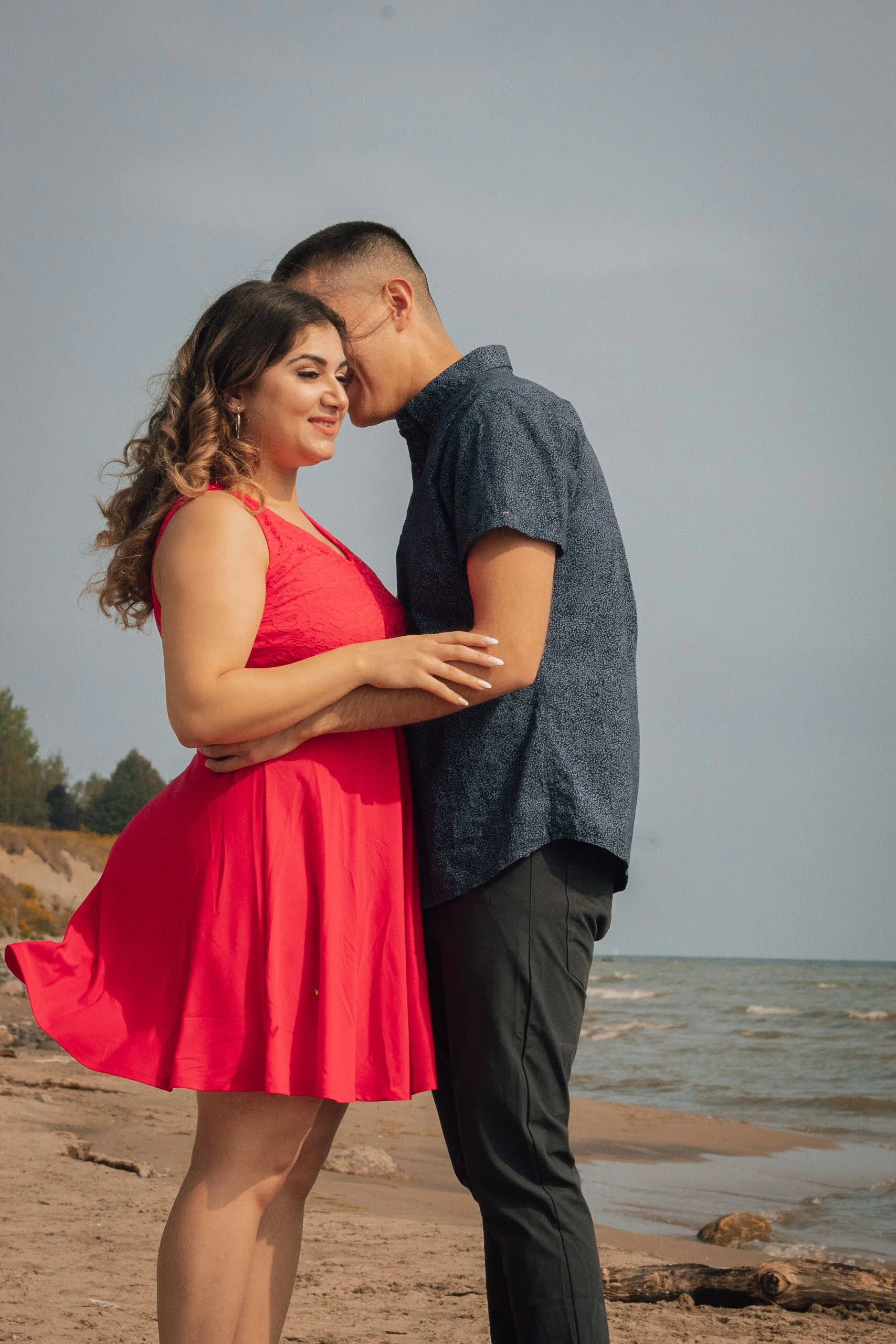 A couple standing close together on a beach, with the woman wearing a red dress and the man dressed in dark pants and a blue shirt, with the ocean in the background.