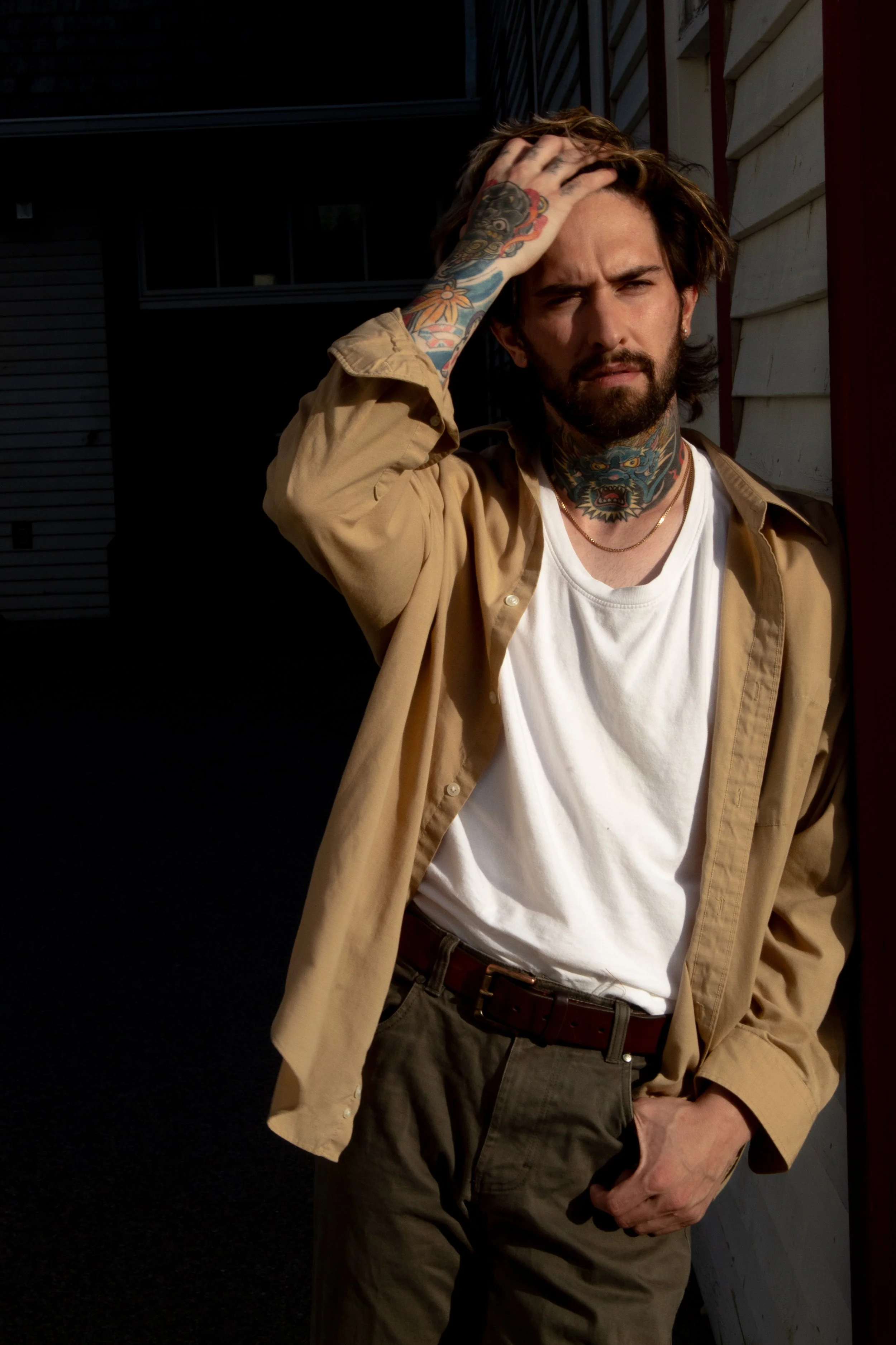 A man with tattoos on his neck and left arm, wearing a beige shirt over a white T-shirt, standing outside near a building with siding. He has dark, shoulder-length hair and a beard, and is holding his head with his left hand, looking into the camera.