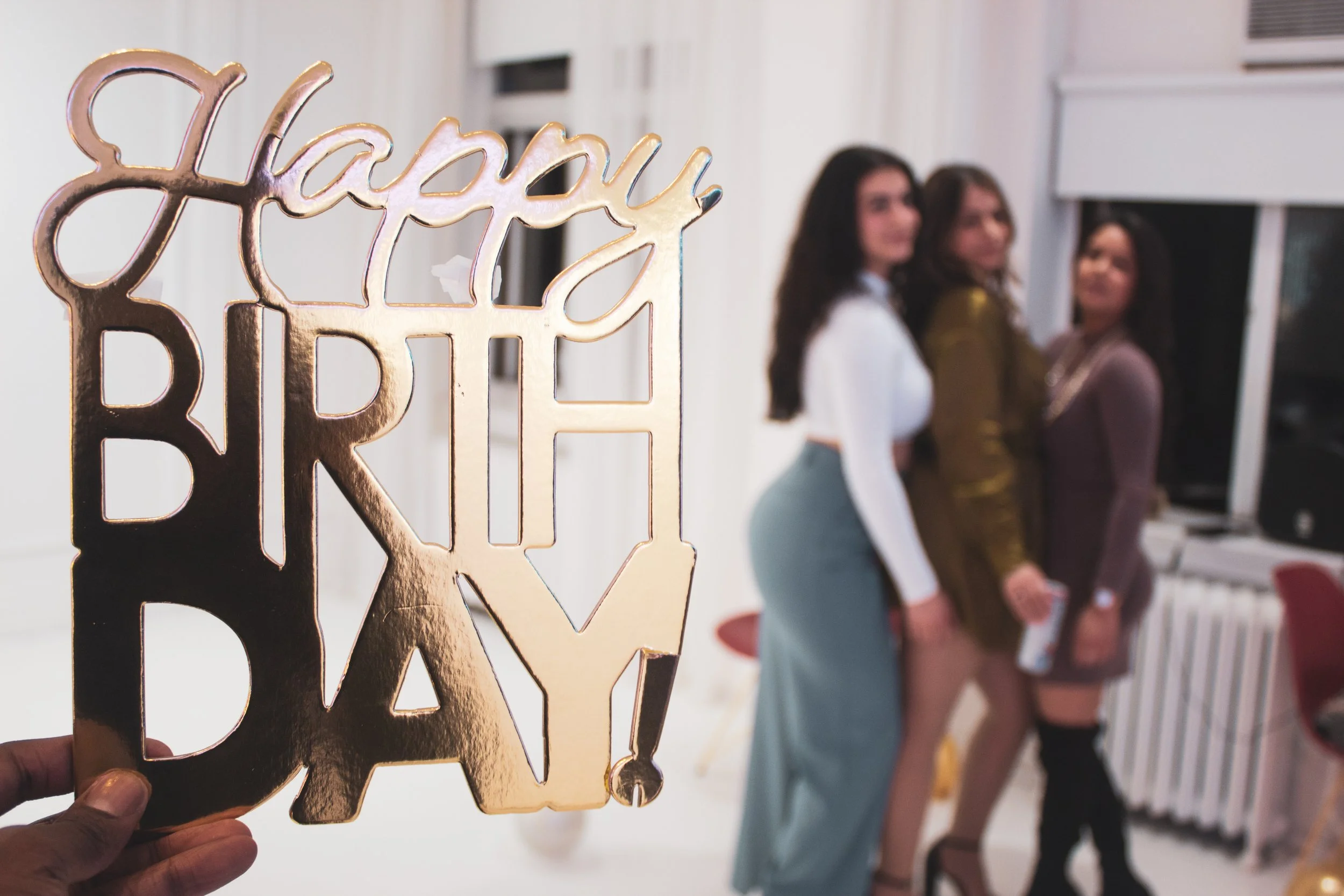 A person holding a gold decorative sign that reads 'happy birthday' in front of three women standing in a line inside a room with white walls and furniture.