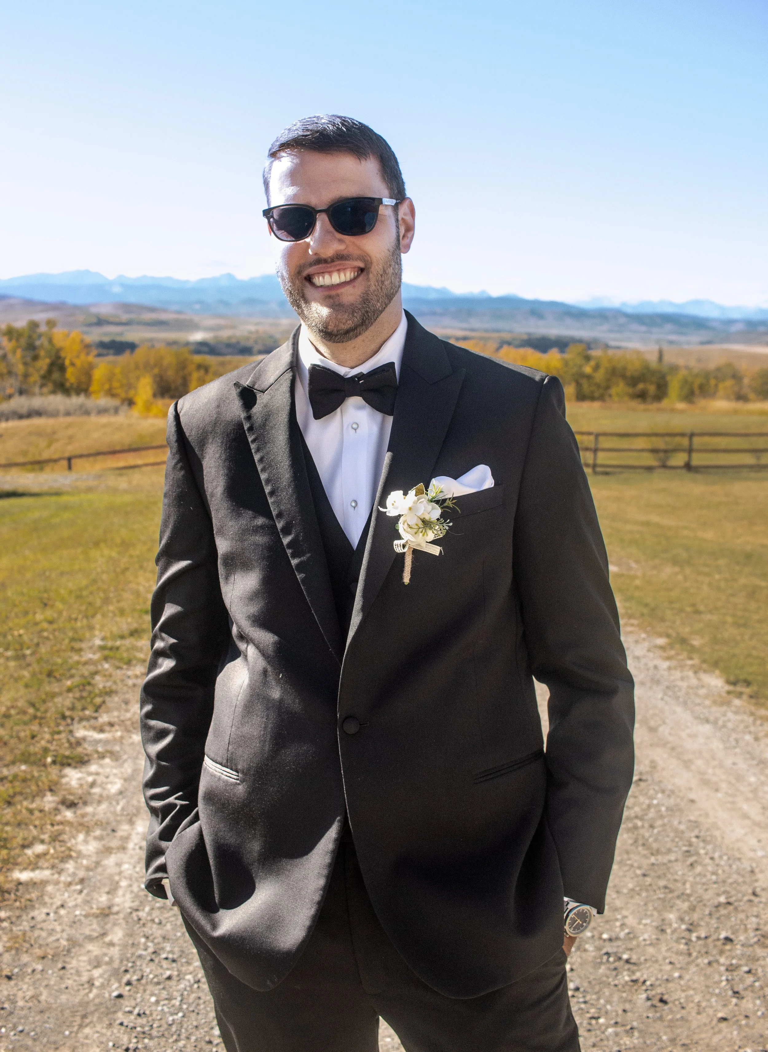 A man in a black tuxedo with a white dress shirt, black bow tie, boutonniere, and sunglasses smiling outdoors on a gravel path with fields, trees, and mountains in the background.