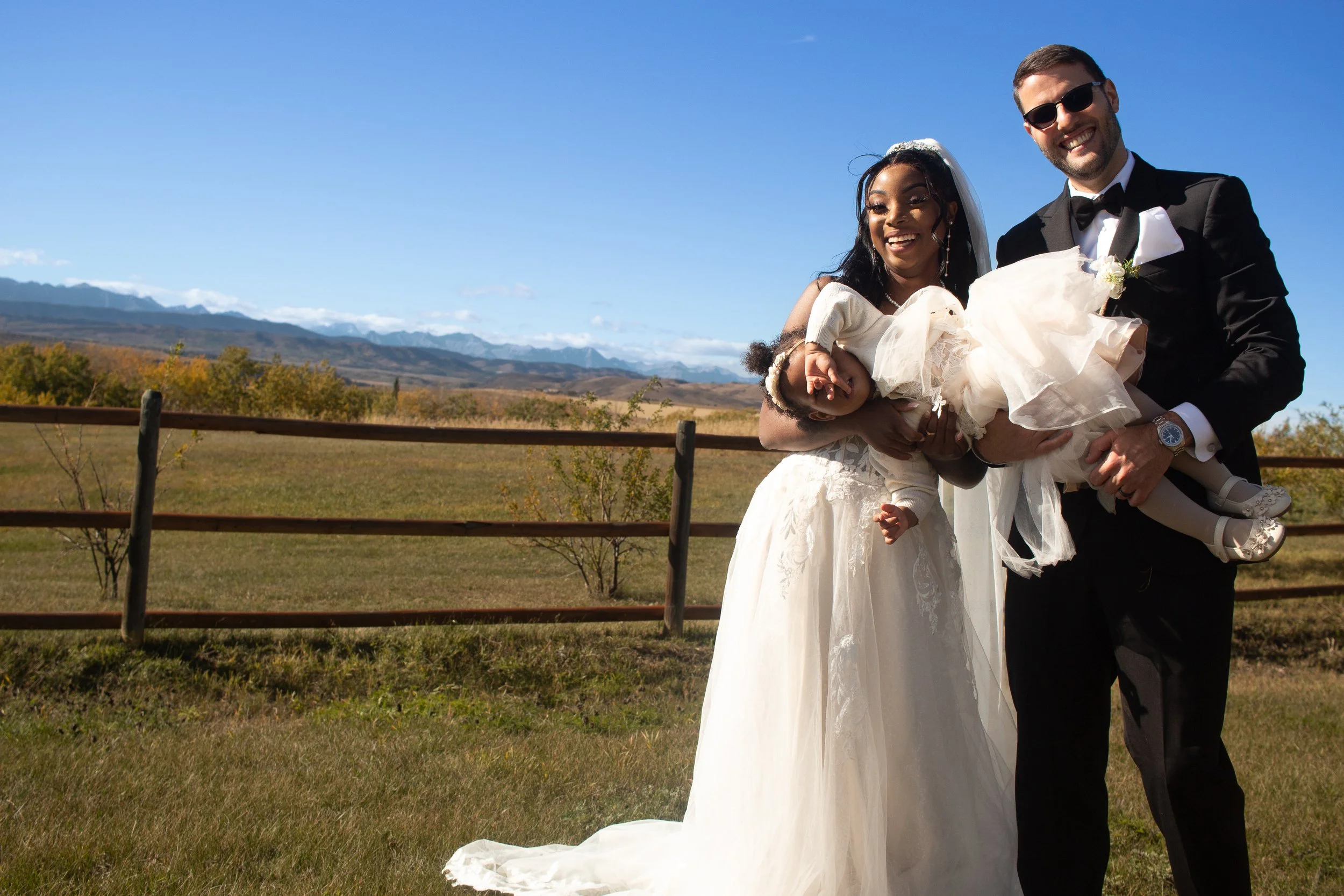 A bride and groom are standing outdoors on a sunny day, with the groom holding a young girl dressed in a white dress. The bride is wearing a white wedding gown and veil, while the groom is dressed in a black tuxedo with sunglasses and a bow tie. They
