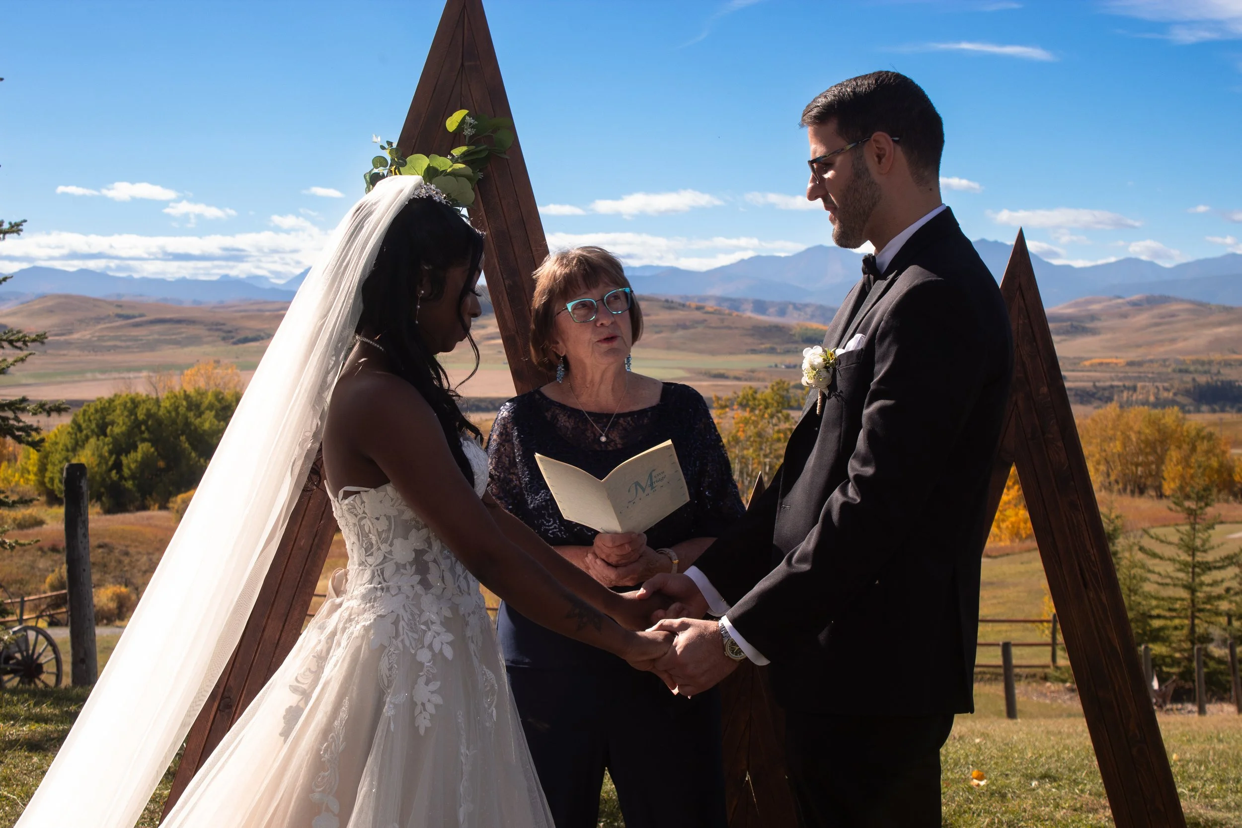 A wedding ceremony outdoors with a bride and groom holding hands while an officiant reads from a book. The bride wears a white lace wedding dress and veil, and the groom wears a black tuxedo. They are under a wooden triangular structure decorated wit