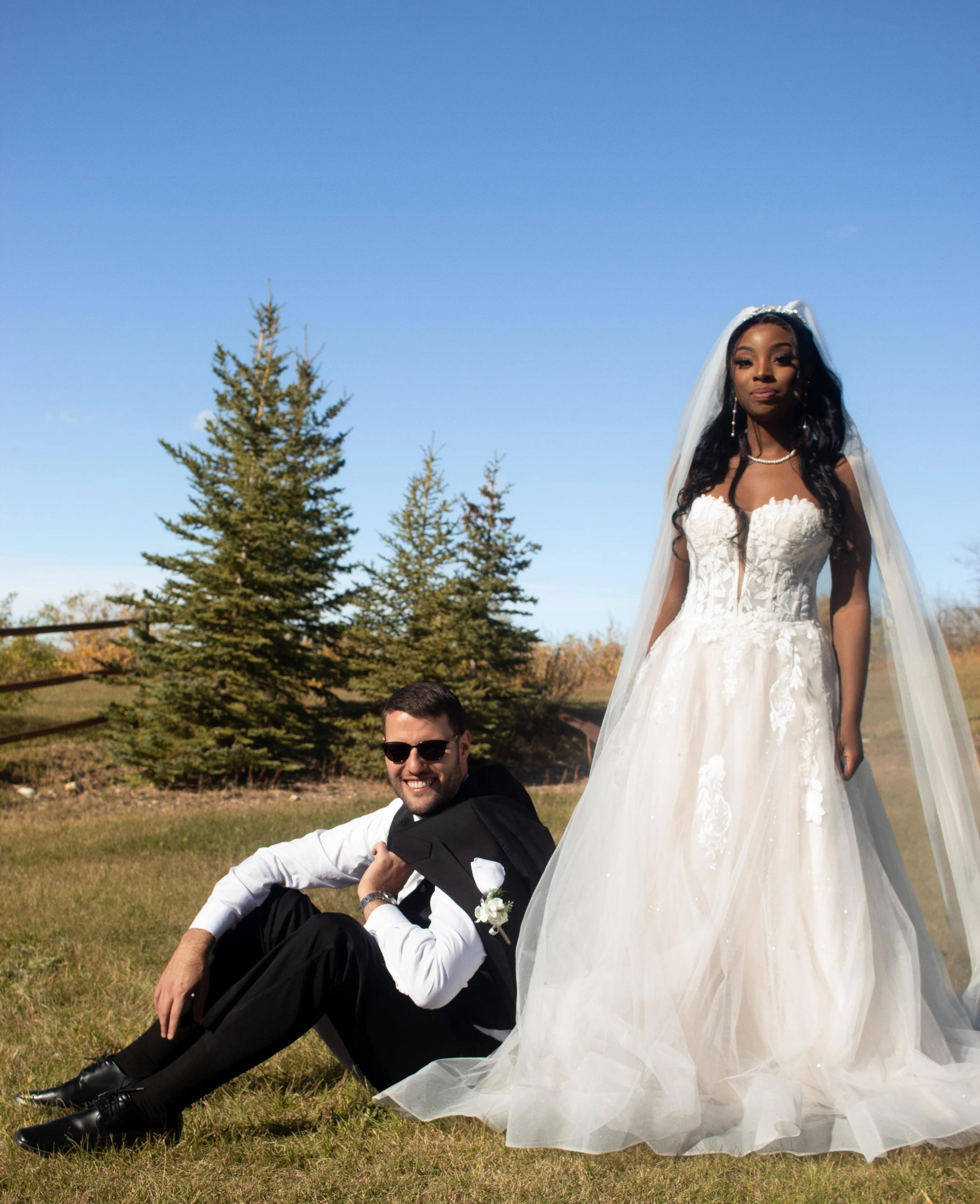 A bride in a white wedding gown and veil standing outdoors, smiling softly, with a man sitting on the grass nearby wearing sunglasses and a tuxedo.