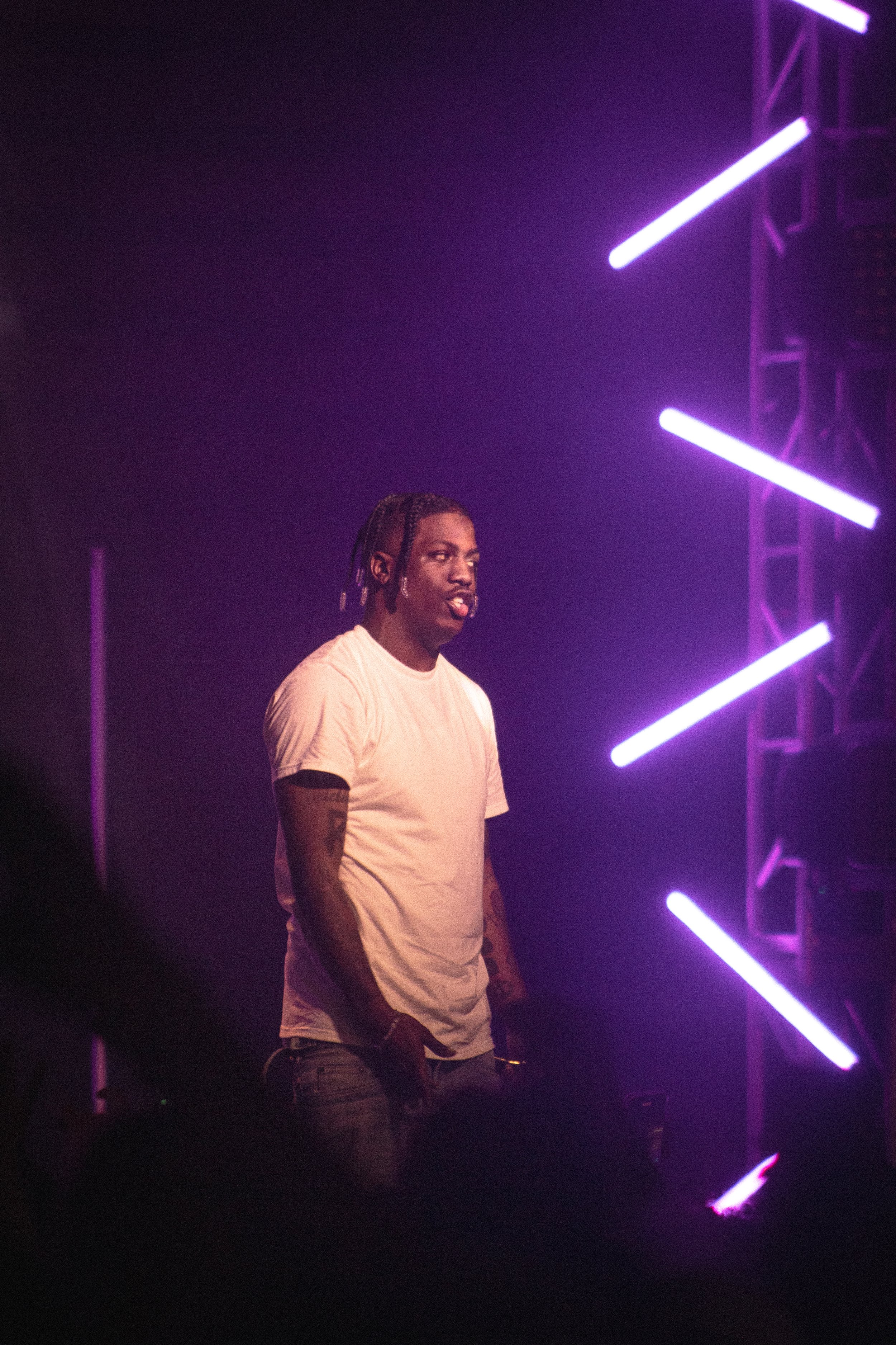 A man with braided hair wearing a white t-shirt stands in front of purple neon lights.