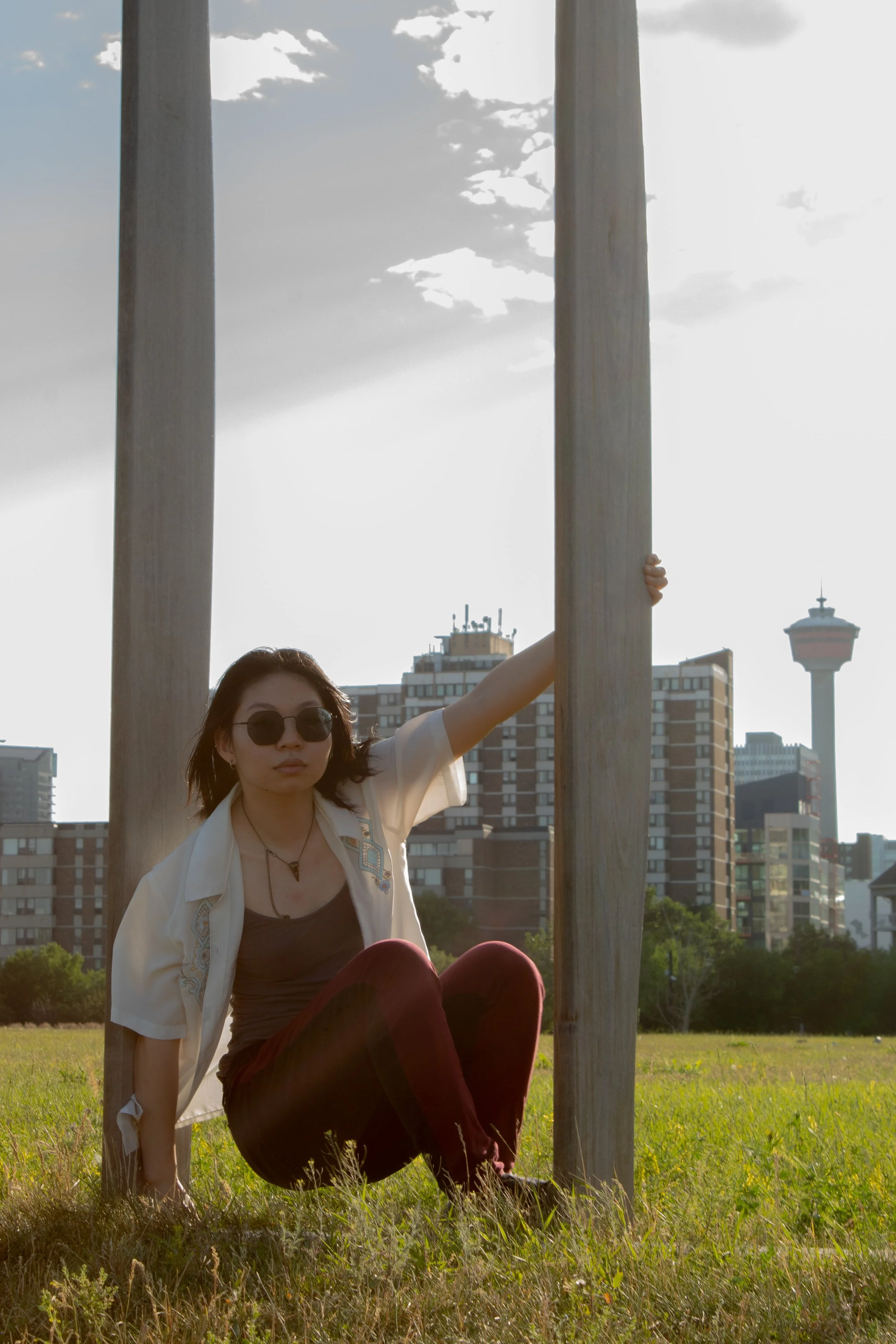 A woman with short dark hair, wearing sunglasses, a beige jacket, and a black top, sits on grass in a park, holding onto a wooden pole with her right hand. The background shows city buildings and a tower with a spire, indicating an urban park setting