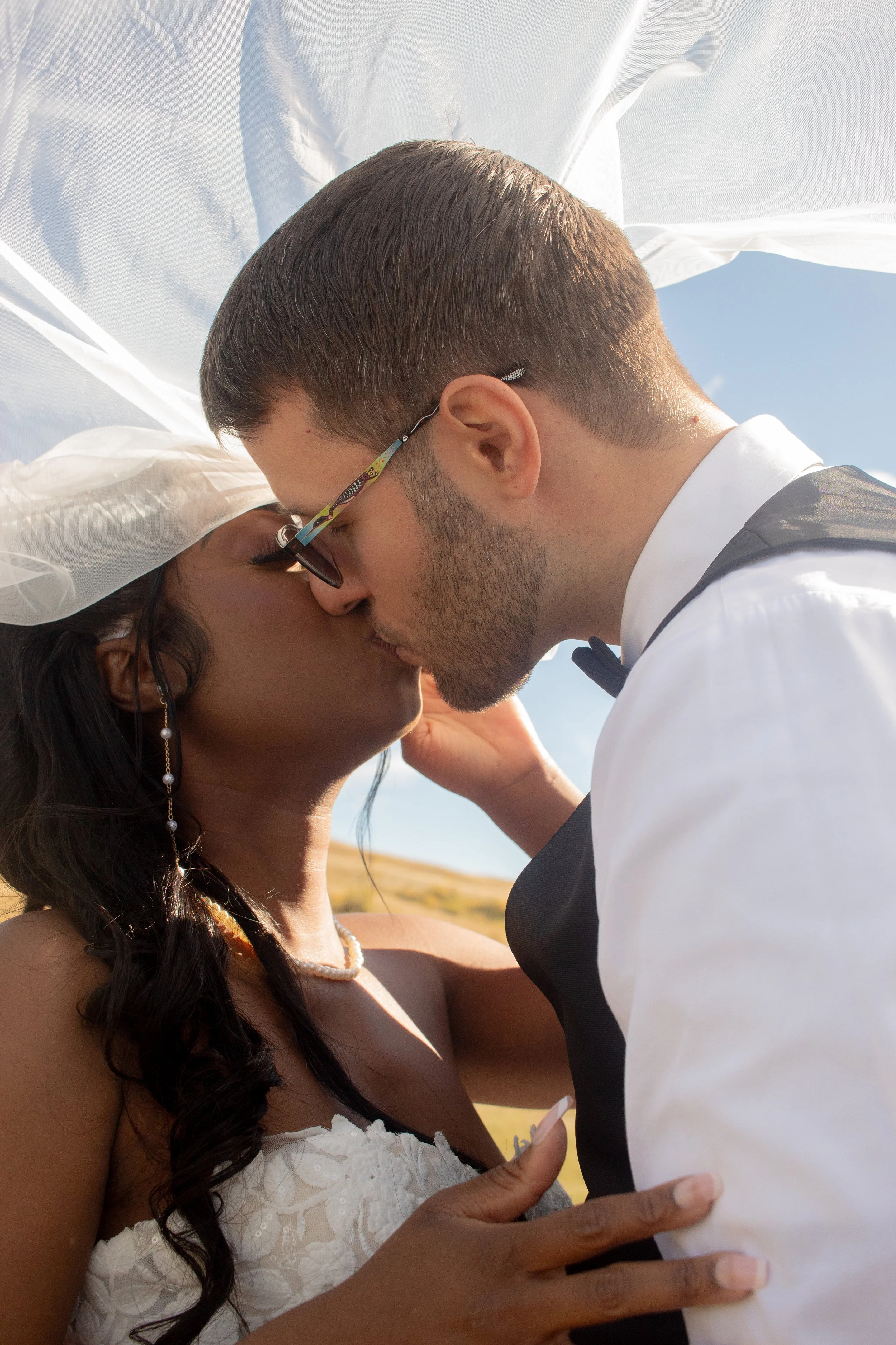 A man and woman share a kiss outdoors under a white canopy, with the woman wearing a white dress and pearl earrings, and the man in a tuxedo.