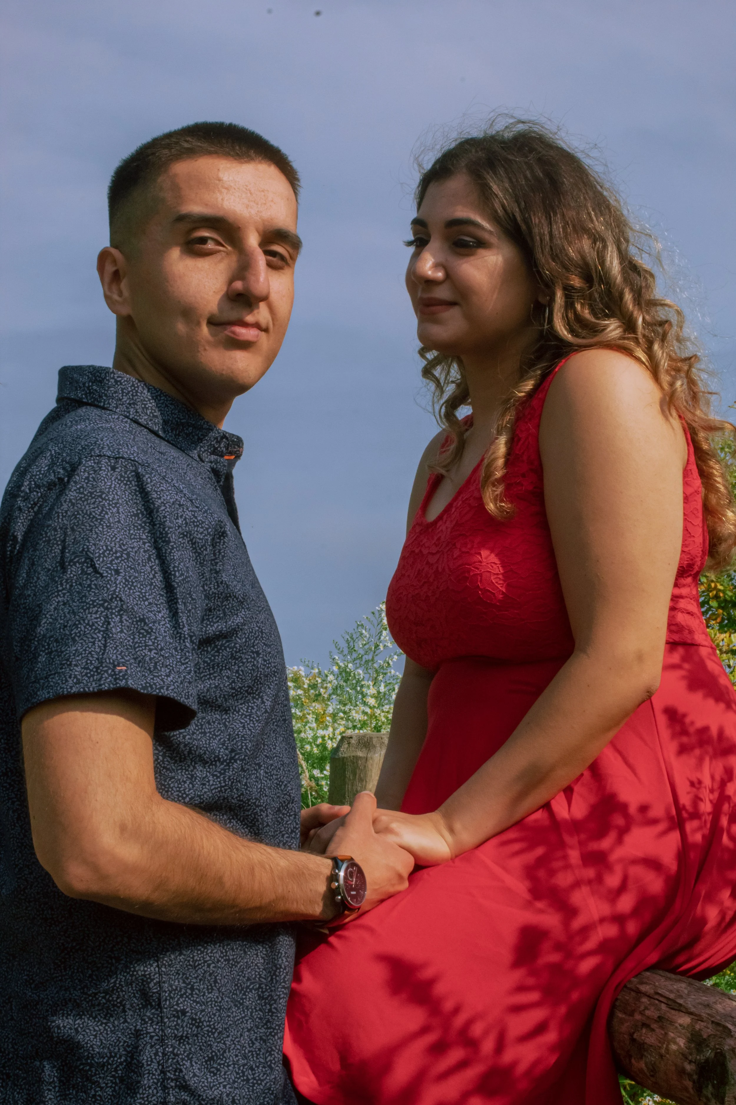 A young man and woman holding hands and facing each other outdoors against a blue sky. The man has short dark hair, wears a patterned dark shirt, and a watch, while the woman has long curly hair, wears a red dress, and is leaning slightly on a tree b