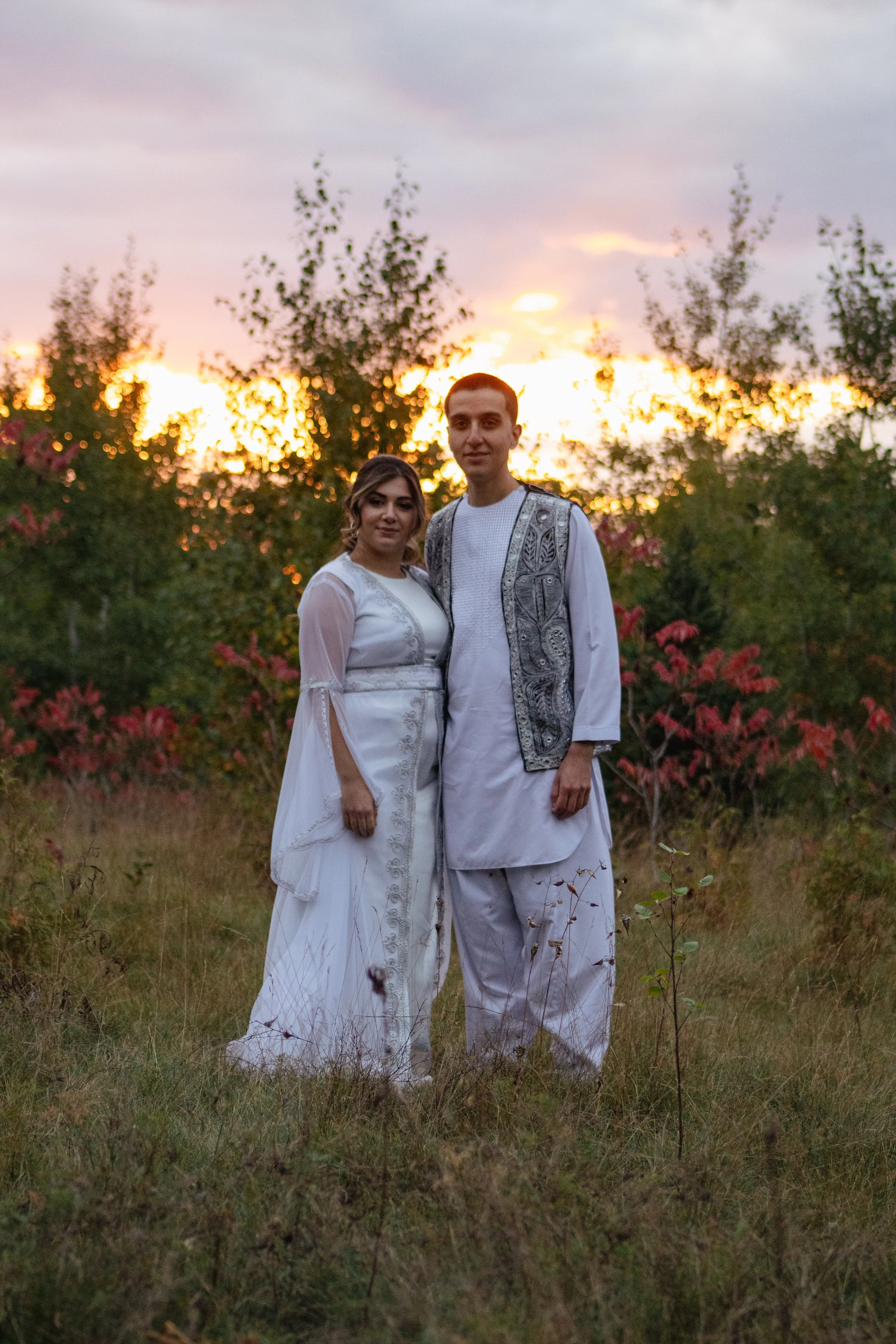 A couple dressed in traditional white attire standing outdoors during sunset, surrounded by trees and grass.