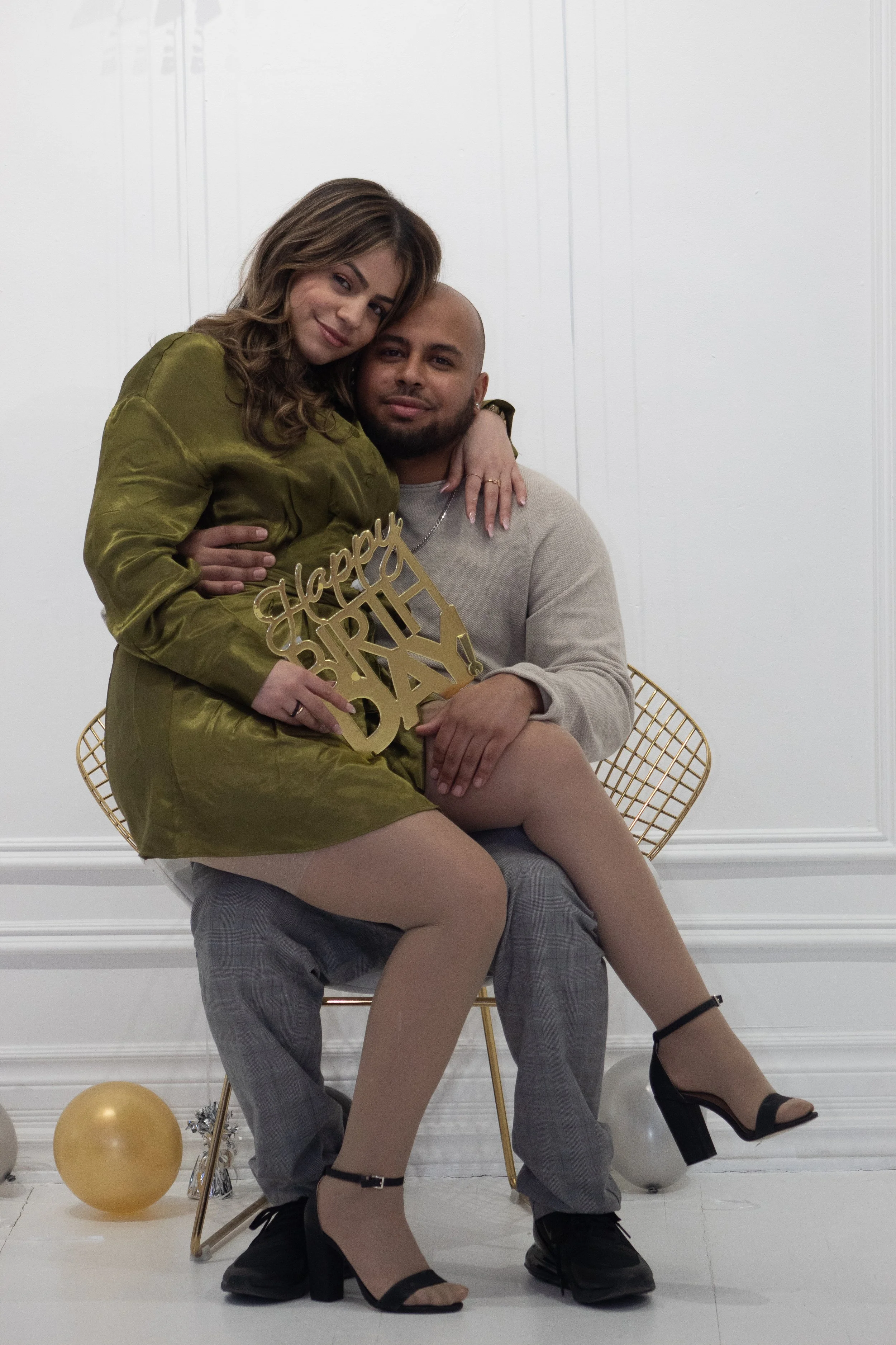 A couple celebrating a birthday indoors with gold balloons and party decorations. The woman is sitting on the man's lap, holding a 'Happy Birthday' sign. They are both dressed stylishly, with the woman in black heels and the man in plaid pants. The b