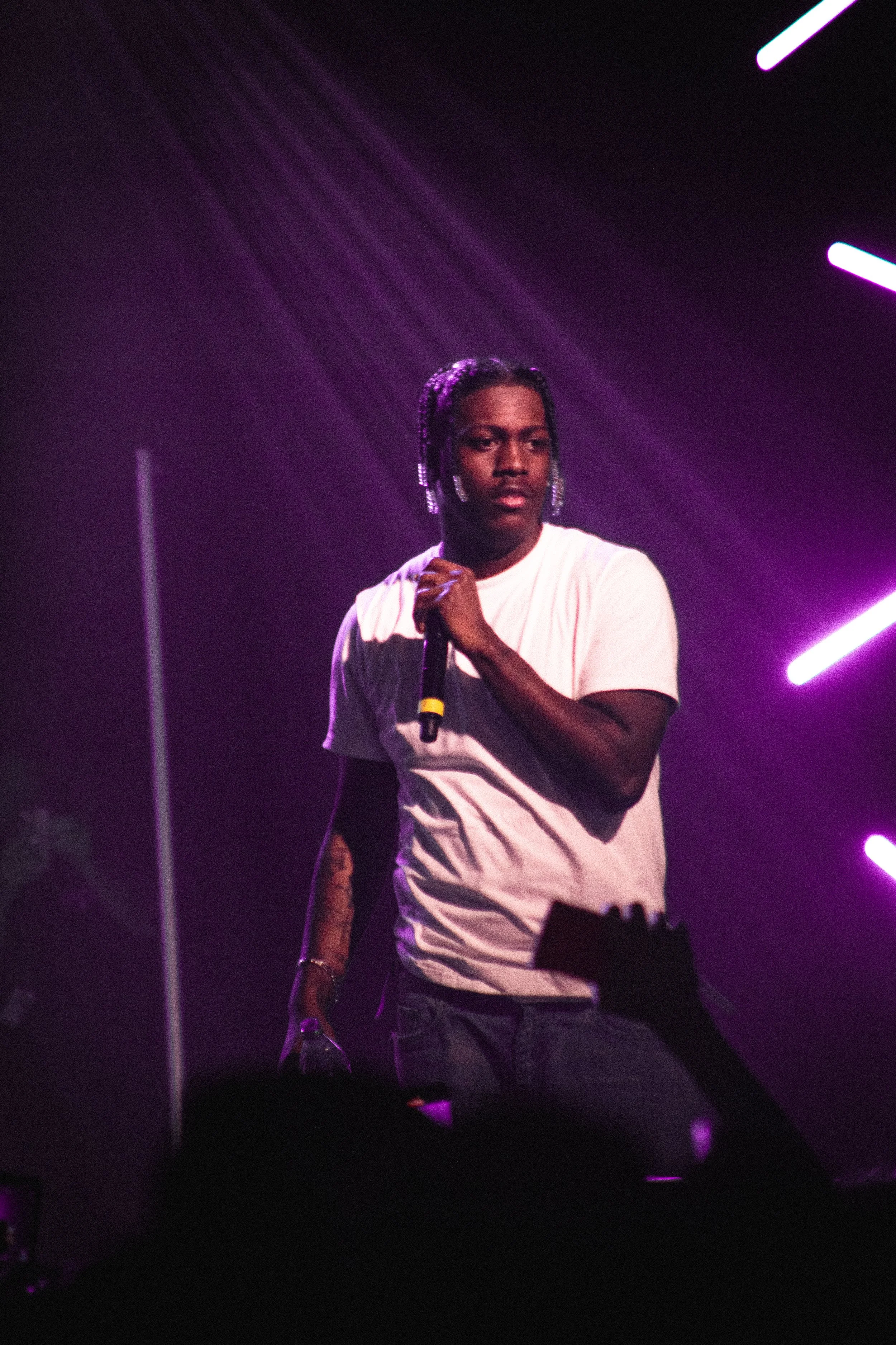 A performer on stage holding a microphone in their right hand, with purple and pink stage lights illuminating the background.