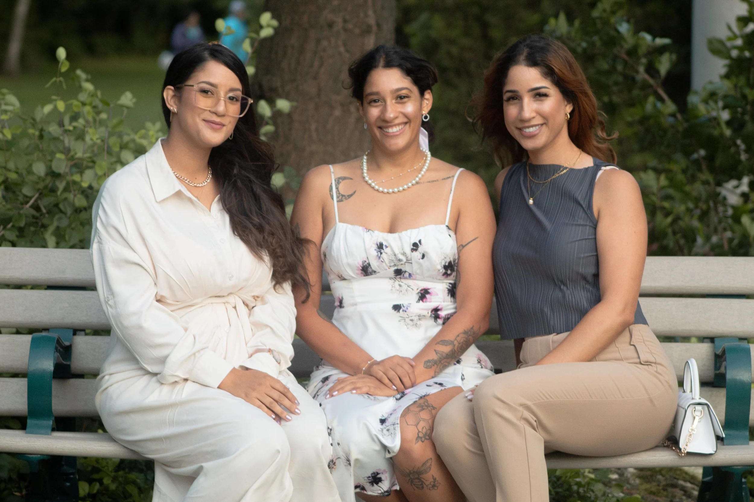 Three women sitting on a park bench, smiling and posing as the camera takes their picture. They are outdoors with green trees and bushes in the background.