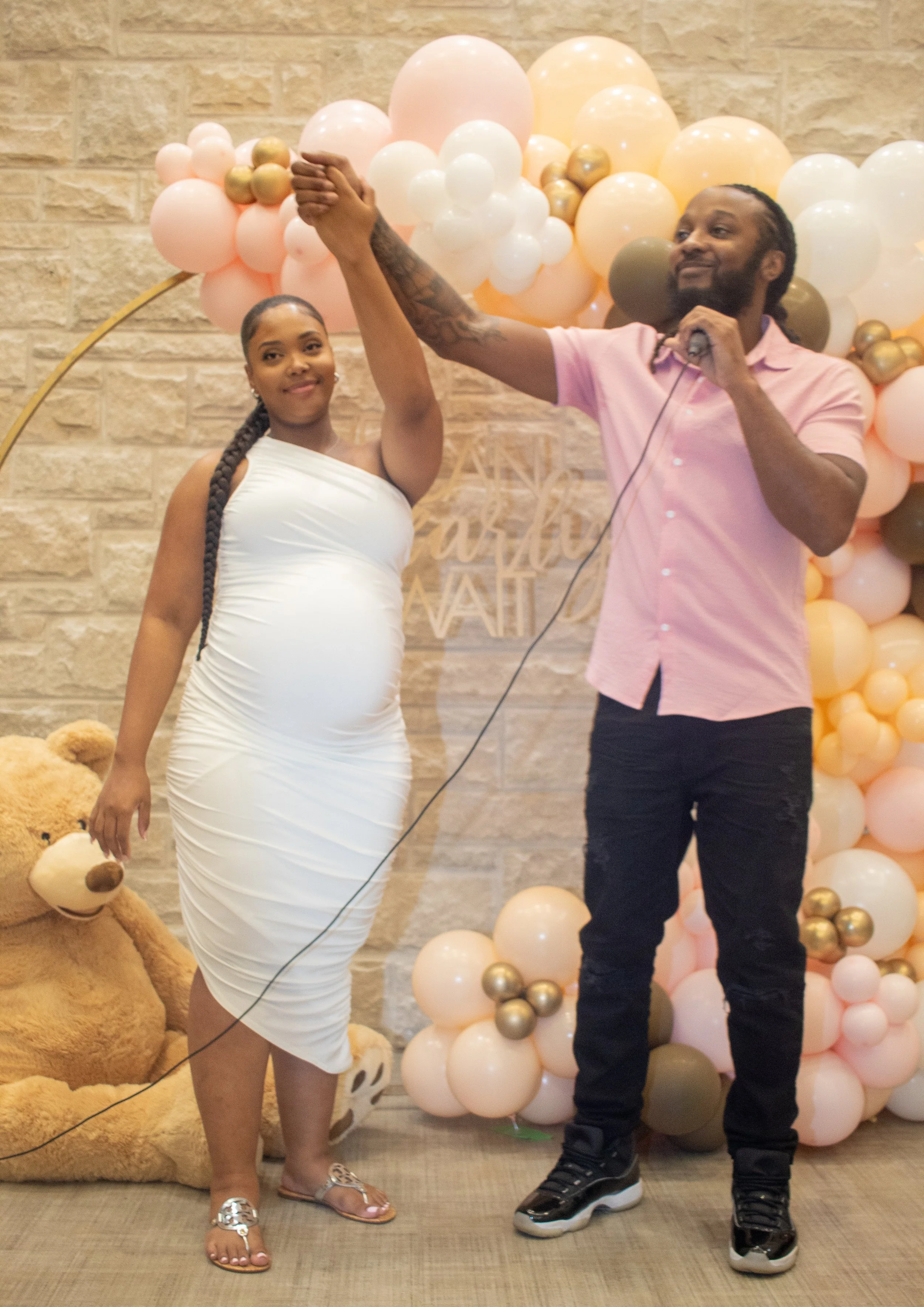 A woman in a white dress and sandals and a man in a pink shirt and black pants are celebrating a baby shower with balloons and teddy bears. They are holding hands in a choreographed pose, with the woman smiling and the man resting his hand on his che