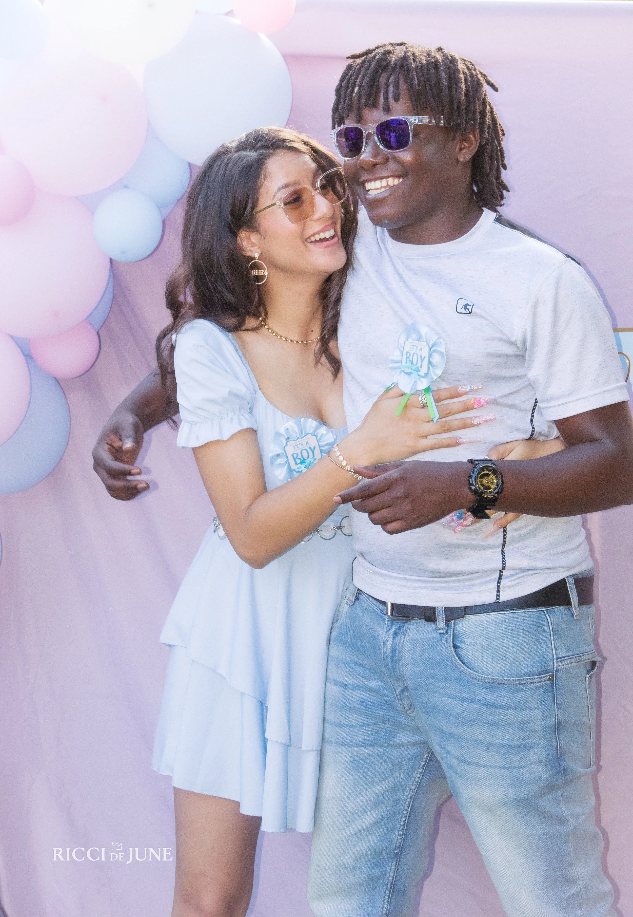 A woman and a man smiling, standing closely together, with balloons in pastel colors in the background. The woman is wearing a light blue dress, glasses, and jewelry. The man is wearing a white T-shirt, sunglasses, and a watch. Many of their accessor