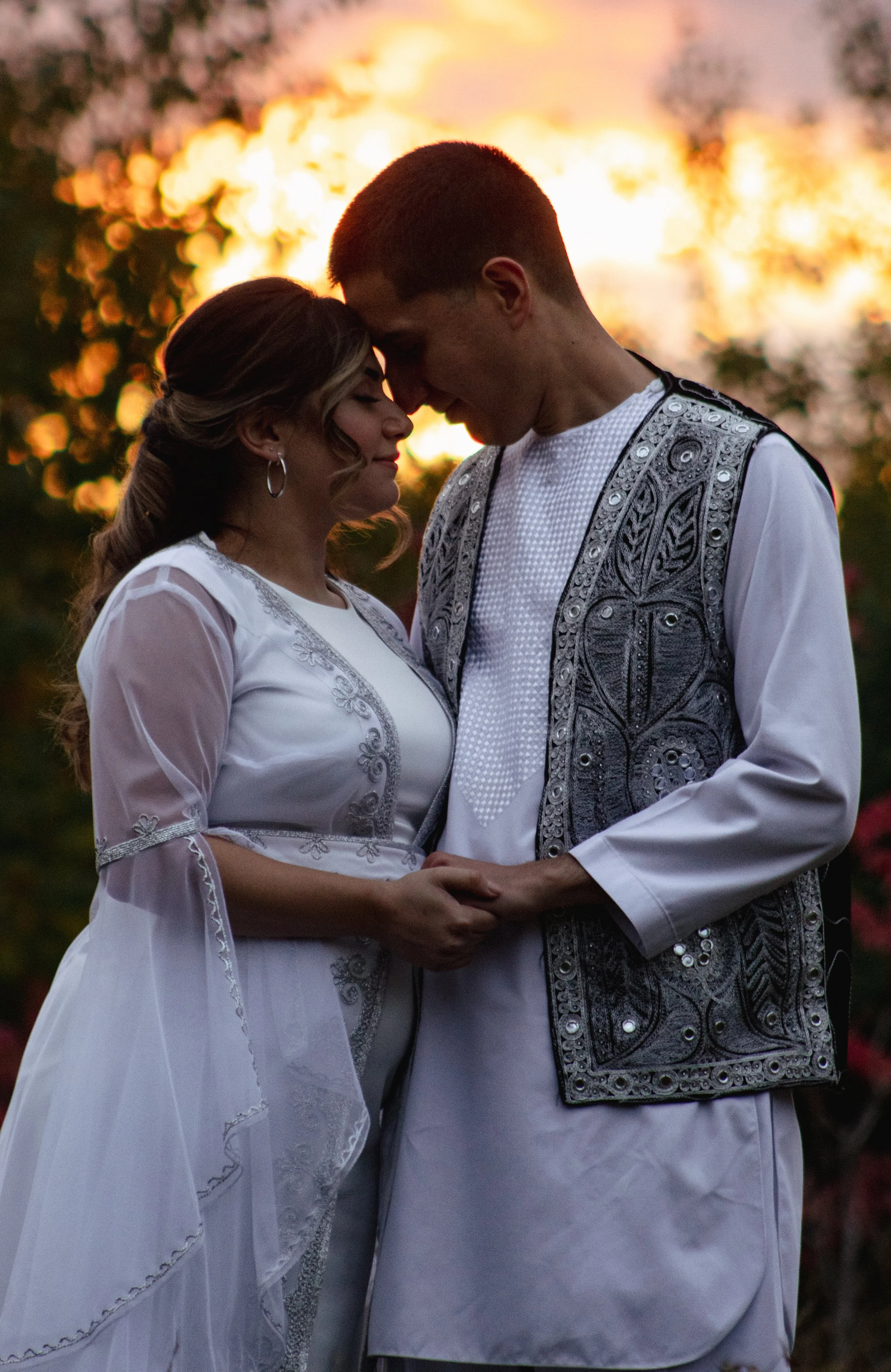 A couple dressed in traditional white and embroidered clothing, holding hands, with pink and orange sunset background.