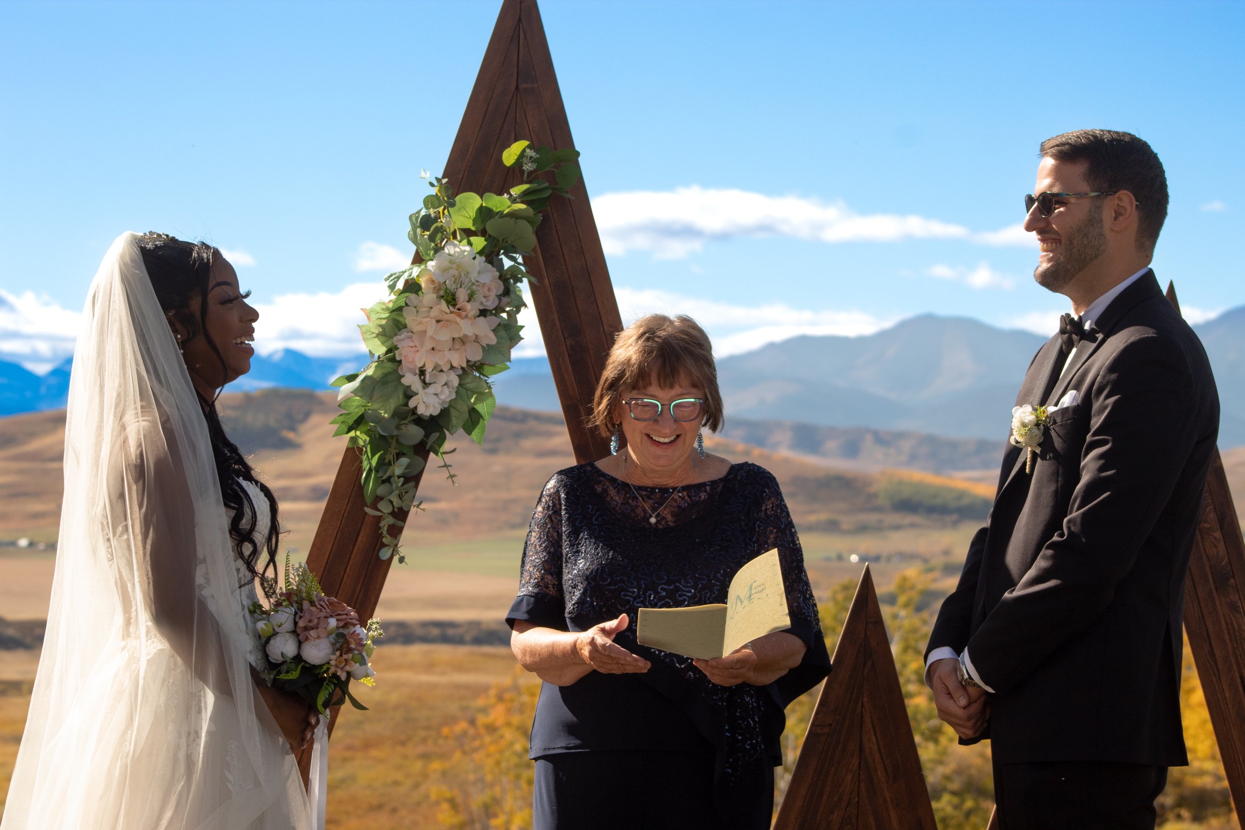 A bride and groom standing during a wedding ceremony outdoors, with an officiant reading from a book. The bride is wearing a white wedding dress and veil, and the groom is in a black tuxedo. There are mountains and blue sky in the background, and a f