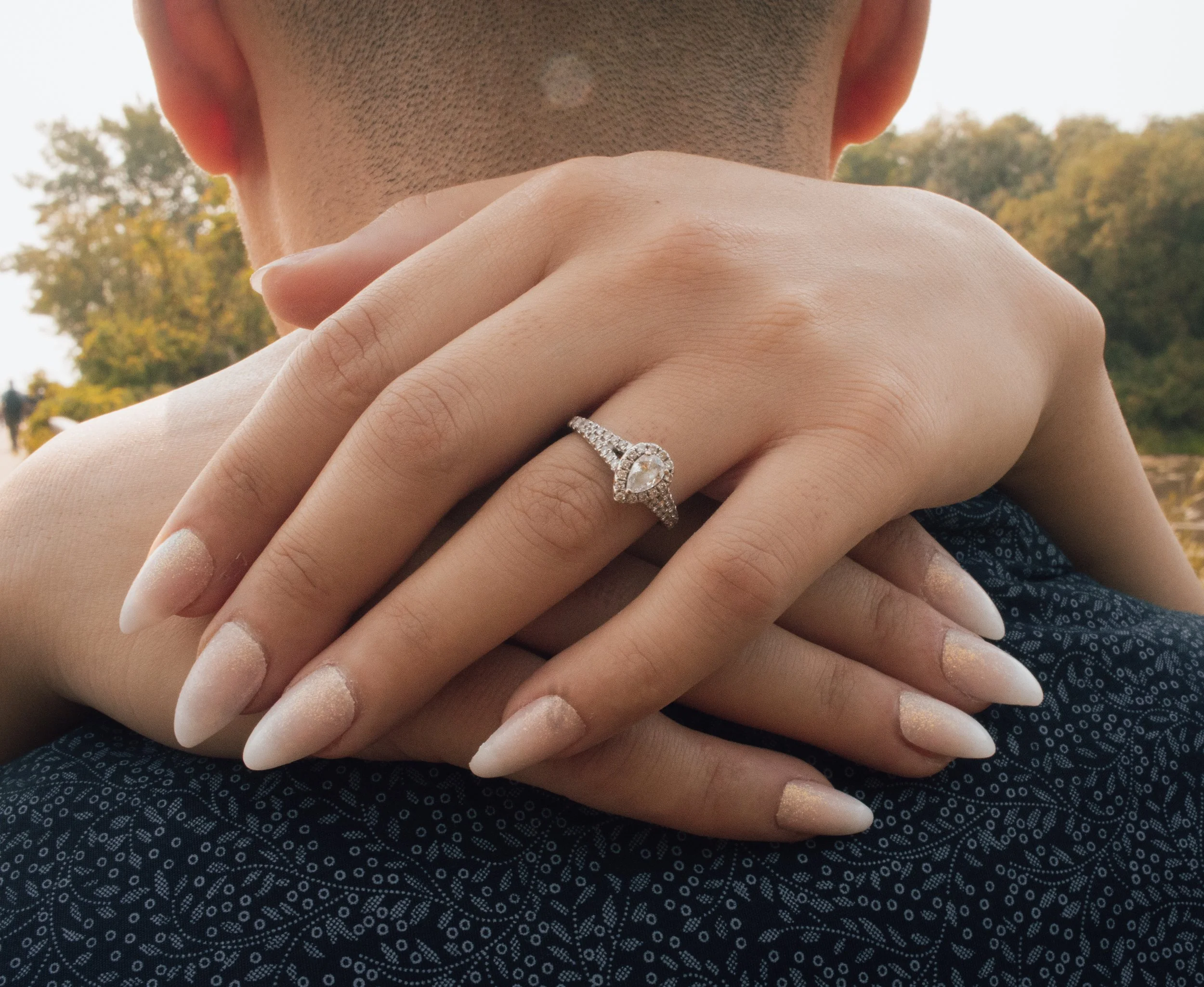 Close-up of two hands, one with a ring, gently holding the neck of another person. The background shows trees with autumn leaves.
