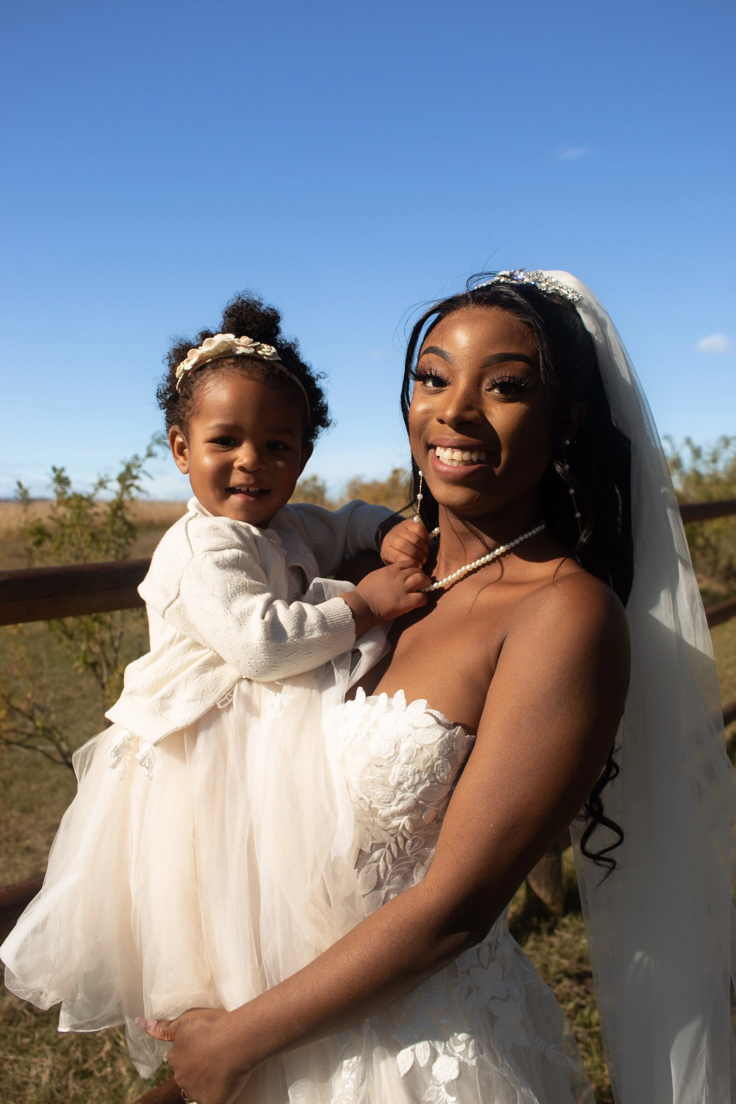 A woman wearing a wedding dress and veil is holding a young girl dressed in a white dress outside under a clear blue sky, with trees and a wooden railing in the background.