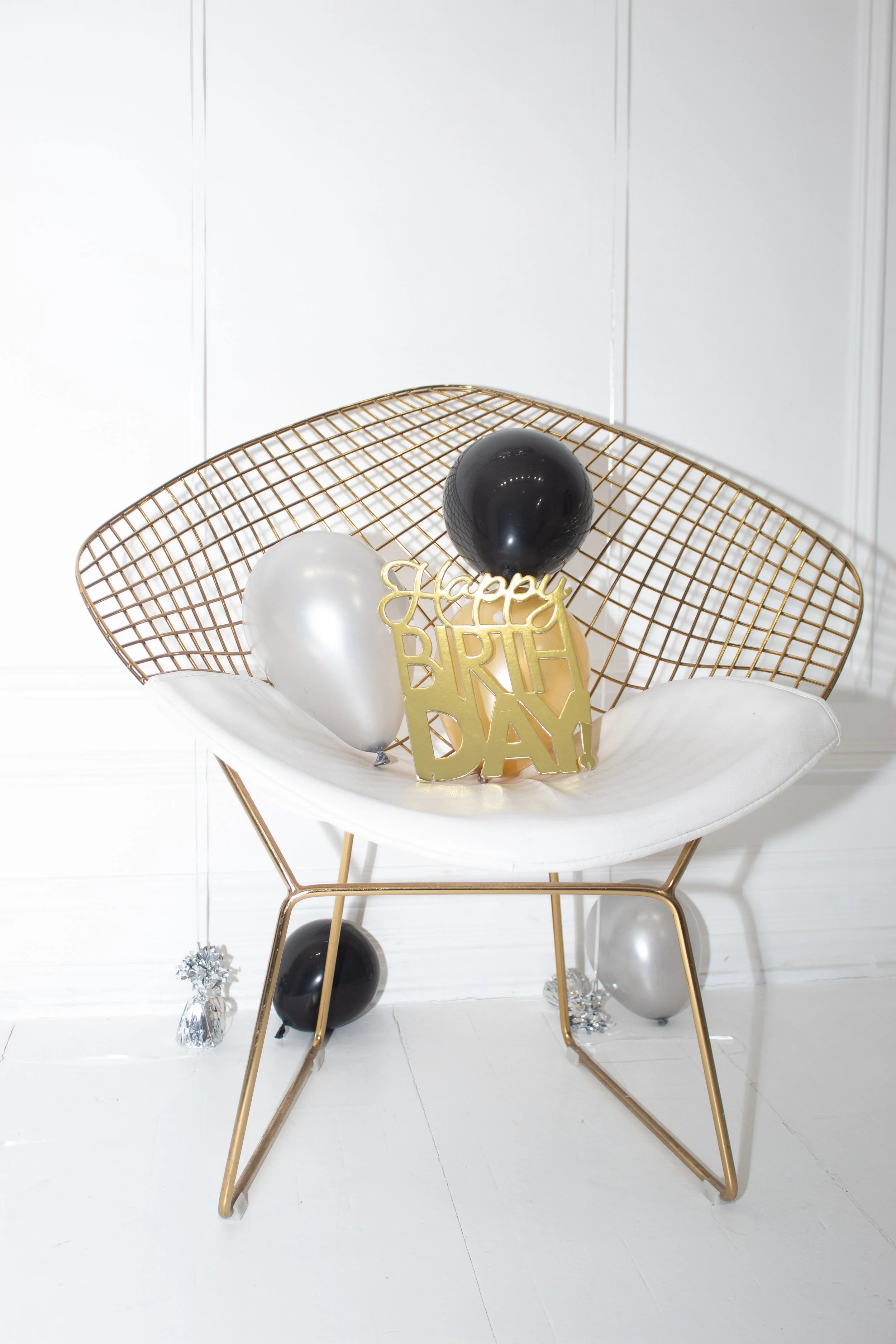 Decorative birthday celebration with black and white balloons, a gold 'Happy Birthday' sign, a gold wire chair, and silver tinsel on a white background.
