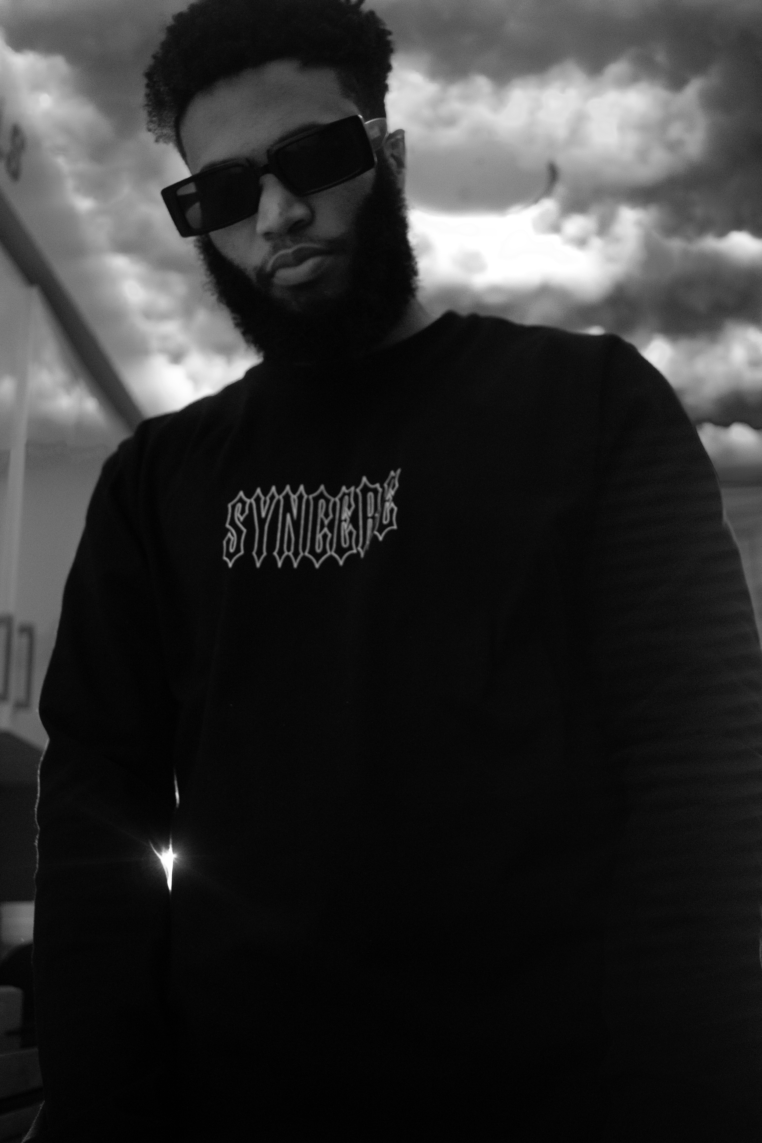 A man with a beard and curly hair wearing large sunglasses and a black t-shirt with a graphic design, standing outdoors against a cloudy sky background.