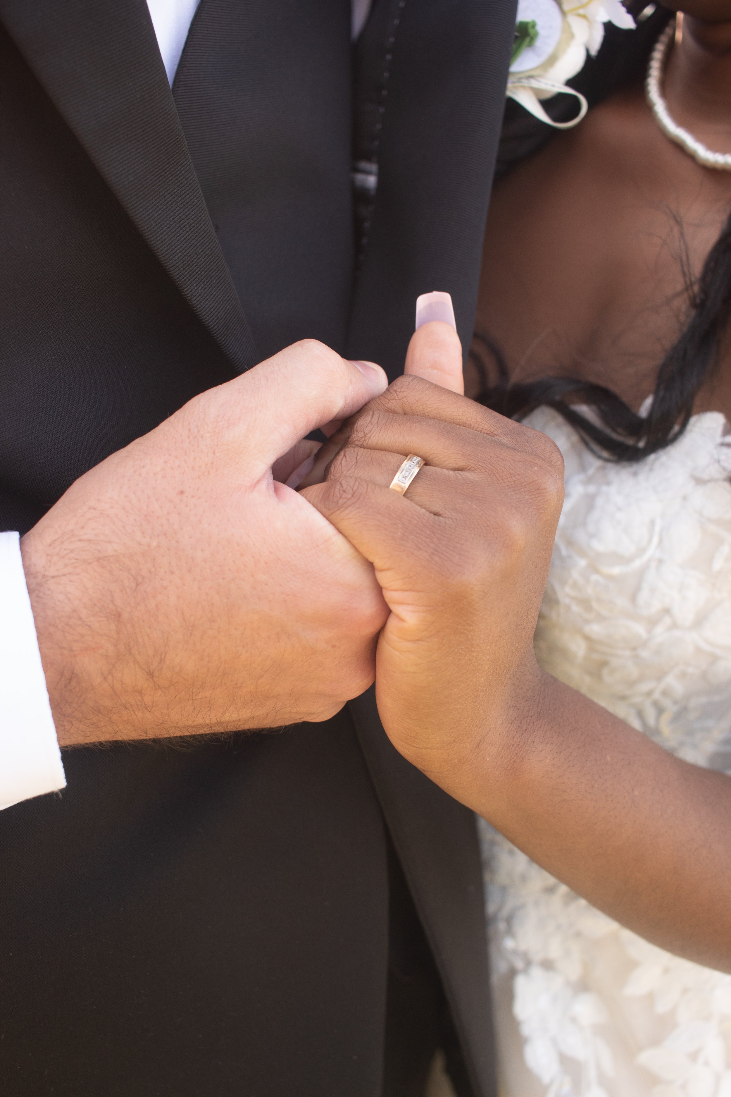Close-up of a bride and groom holding hands during a wedding ceremony, with the groom's hand on top of the bride's hand, showing the wedding ring on the bride's finger. The groom is dressed in a black suit, and the bride is wearing a white lace dress