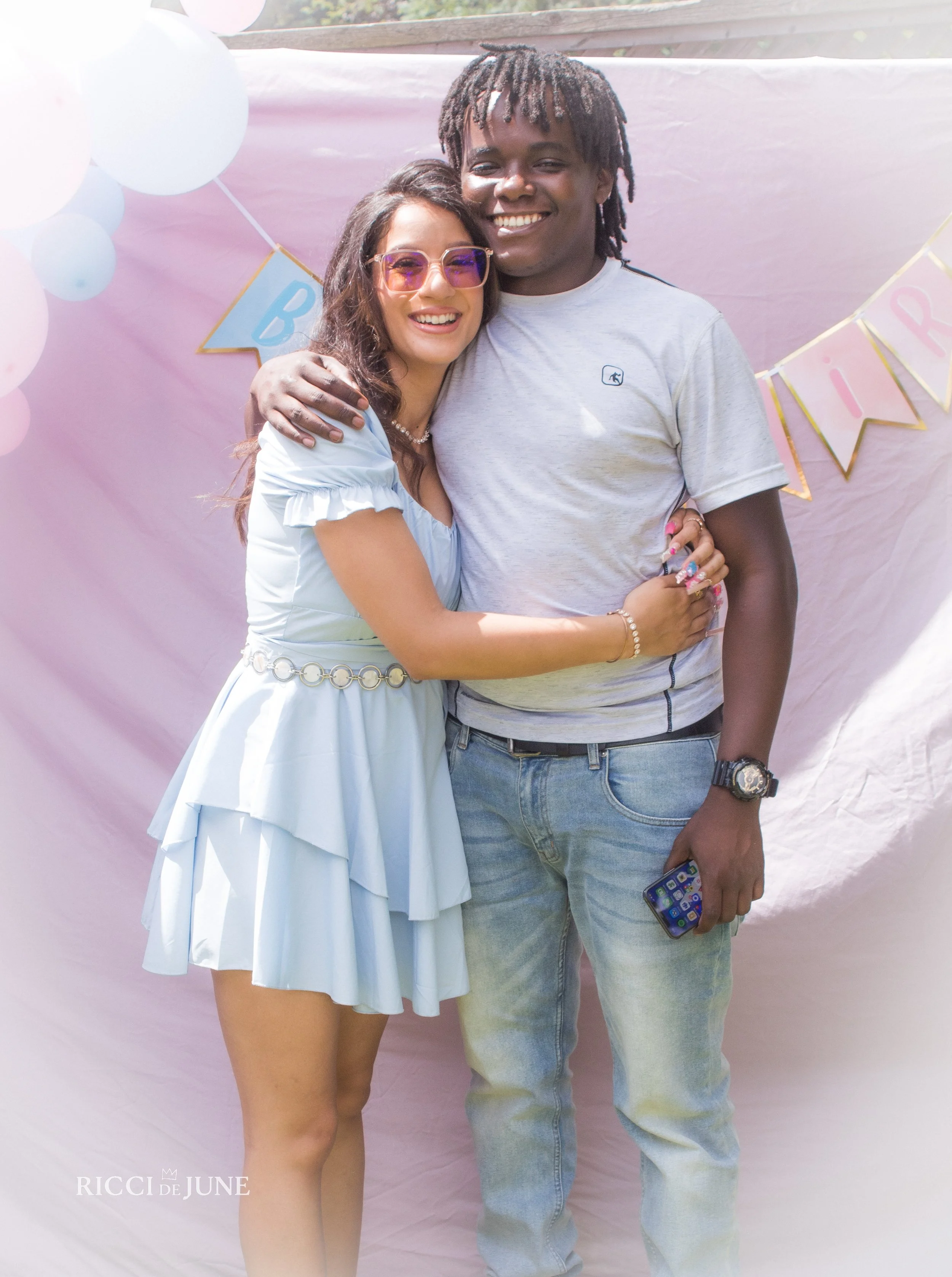 A woman and a man are smiling and hugging in front of a pink backdrop decorated with balloons and a banner. The woman is wearing a light blue dress with ruffles and a pearl necklace. The man is wearing a gray t-shirt and blue jeans, holding a smartph