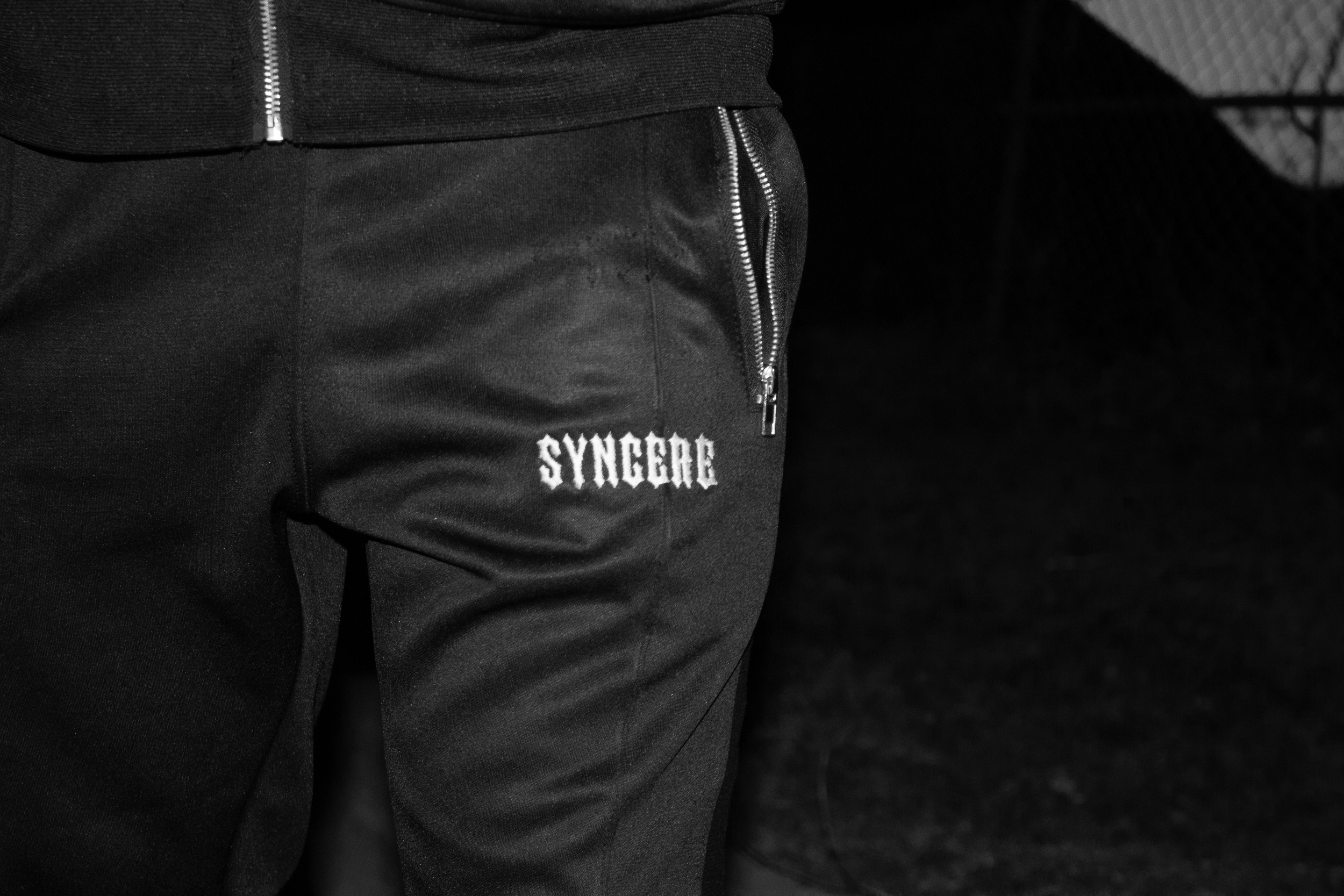Close-up of a person wearing black pants with a zippered pocket and the word 'SYNGER' embroidered on it, part of a black zip-up jacket, and a dark background.