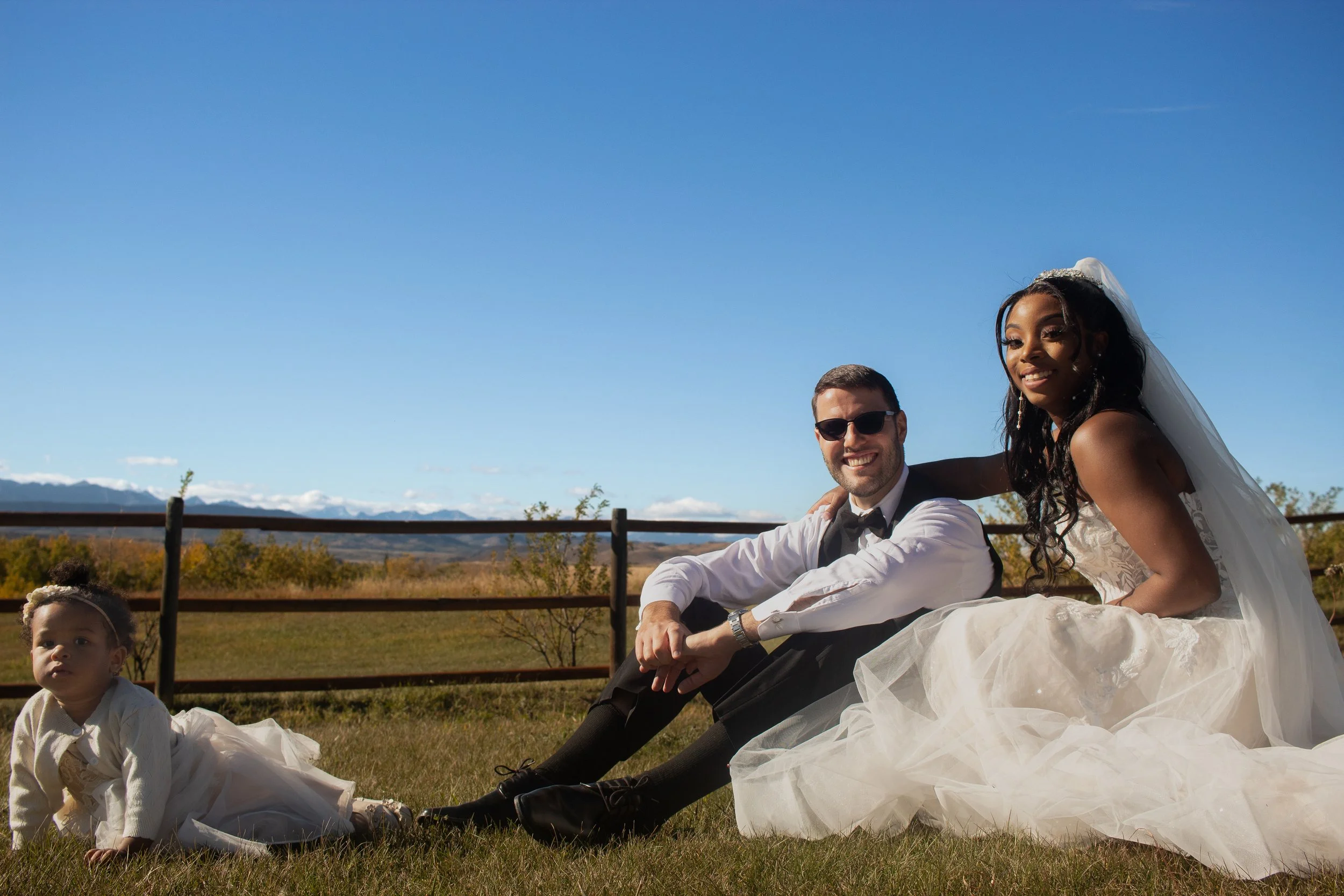 A wedding scene outdoors with a bride and groom sitting on the grass, smiling at the camera, with a young girl nearby. The bride is wearing a white lace wedding dress and veil, and the groom is wearing a white shirt with a bow tie and sunglasses. The
