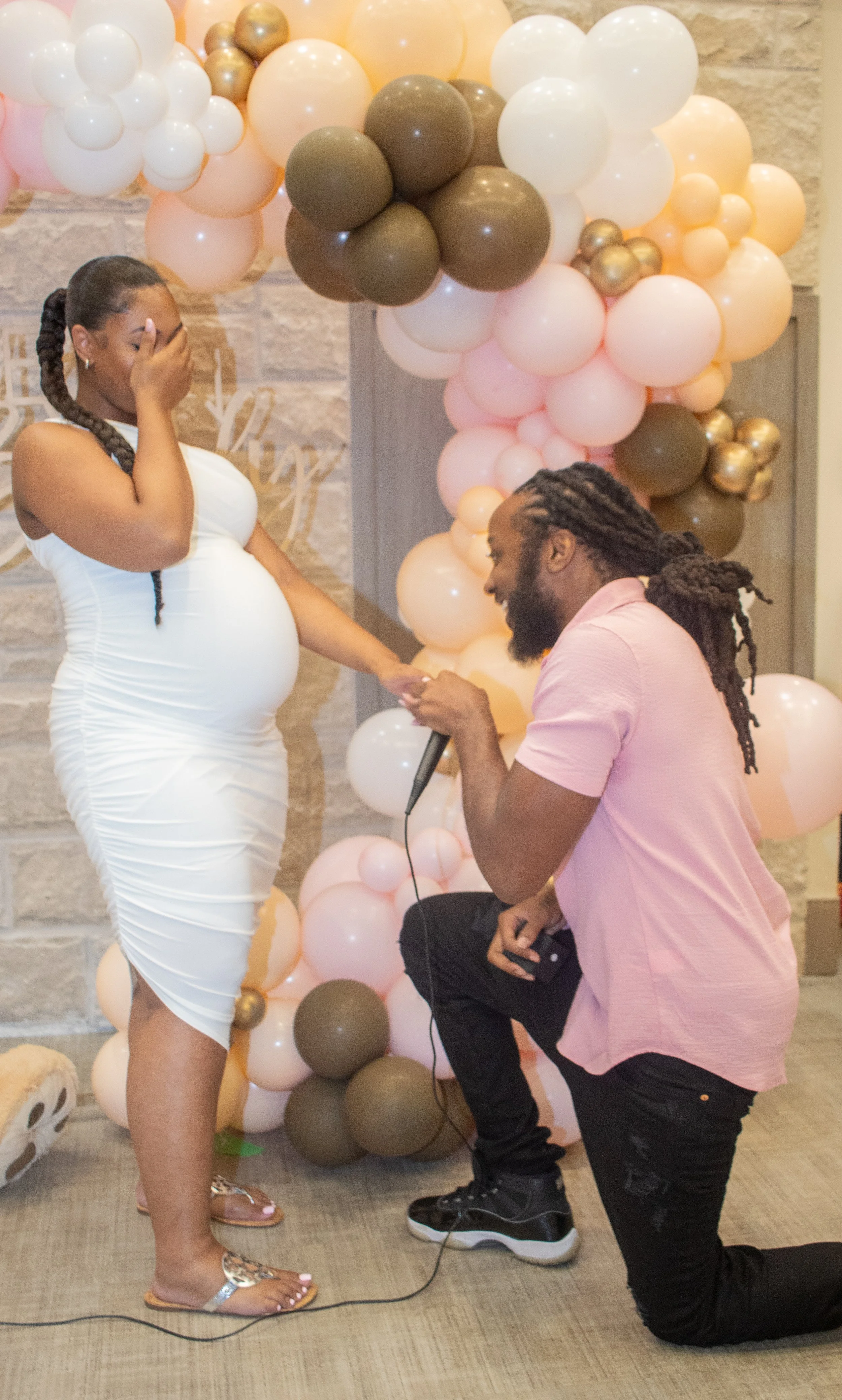 A man is kneeling on one knee, proposing to a pregnant woman in front of a balloon decoration, with a woman covering her face with her hand in surprise.