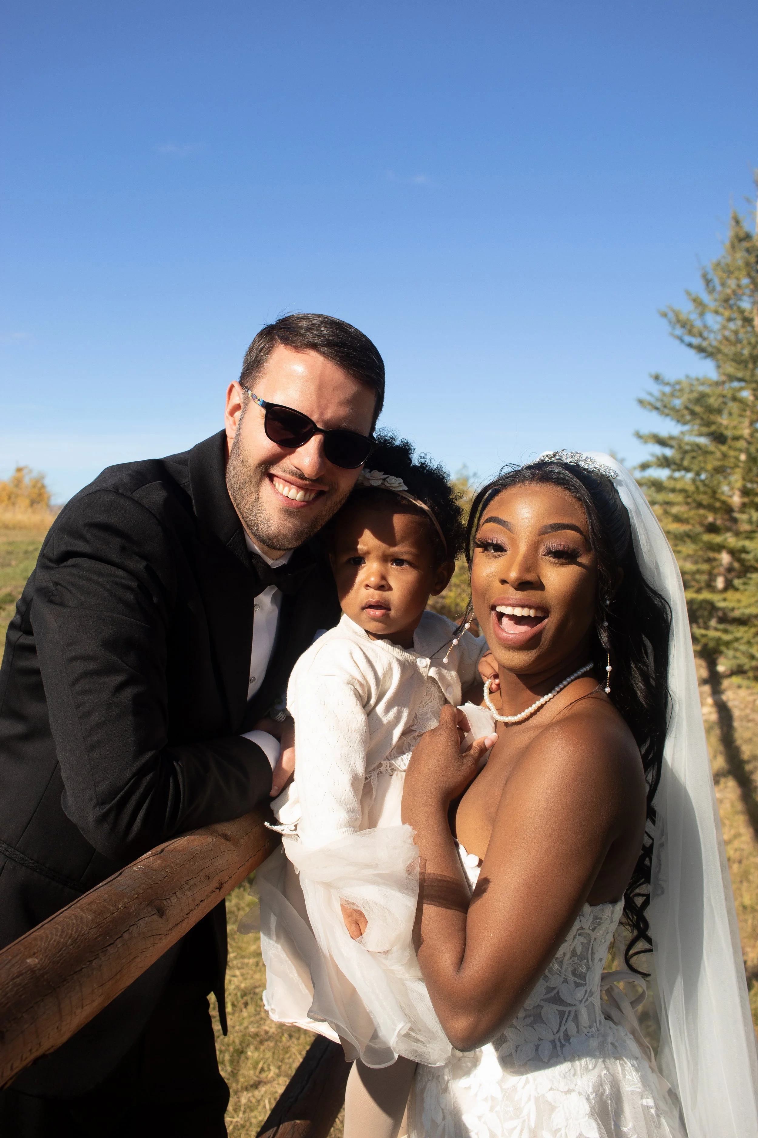 A joyful couple with a young child outdoors on a sunny day, all dressed in formal attire for a wedding, standing by a wooden fence with trees in the background.