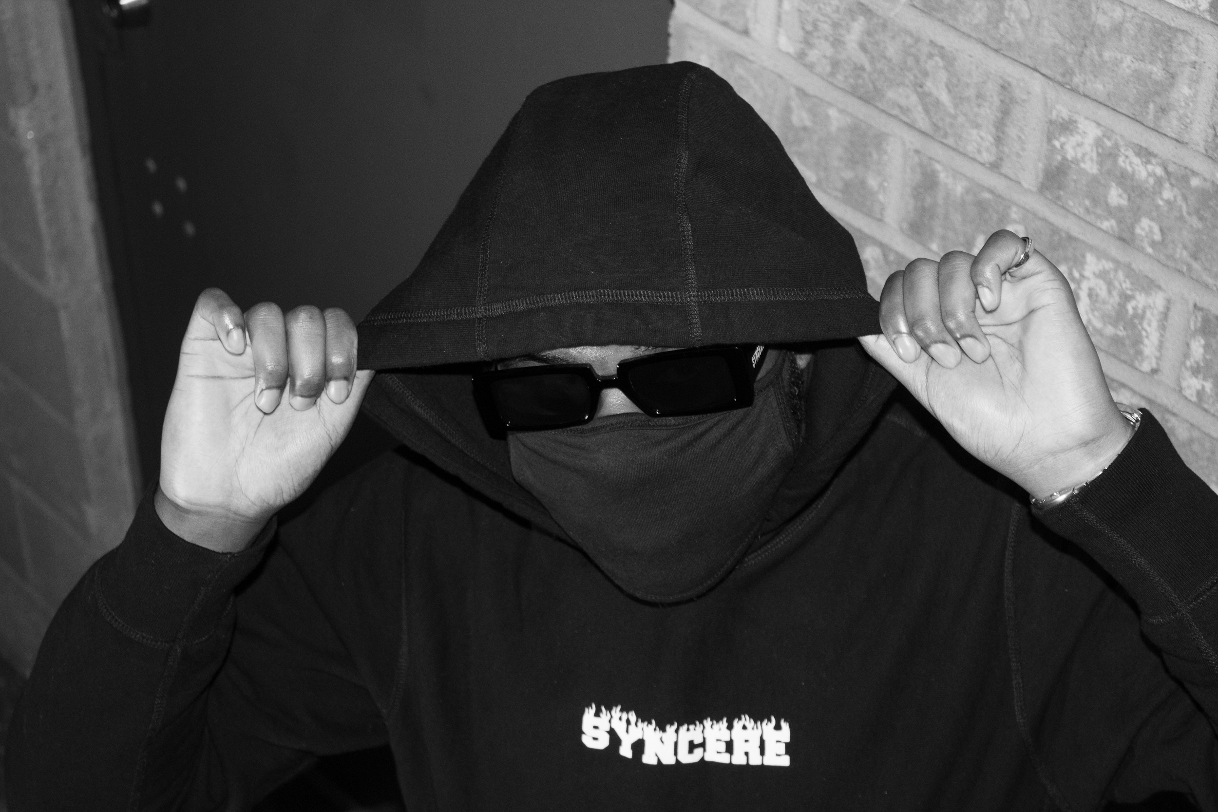 Person wearing a black hoodie, sunglasses, and a shirt with the word 'SINCERE' on it, adjusting their hood, standing against a brick wall and a dark background.
