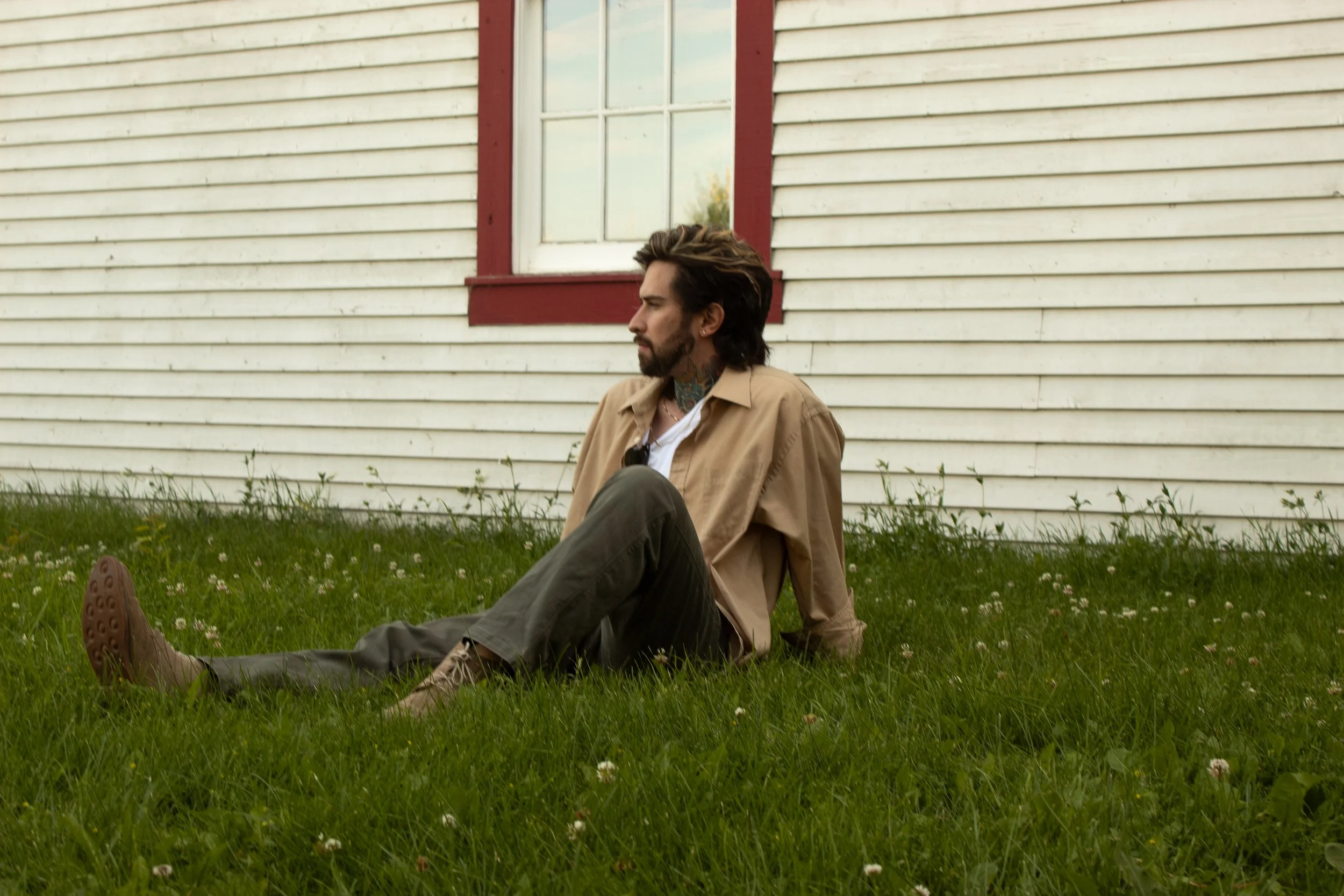 A man with dark hair, beard, and tattoos sitting on grass in front of a white house with red trim around the window, looking to the left.