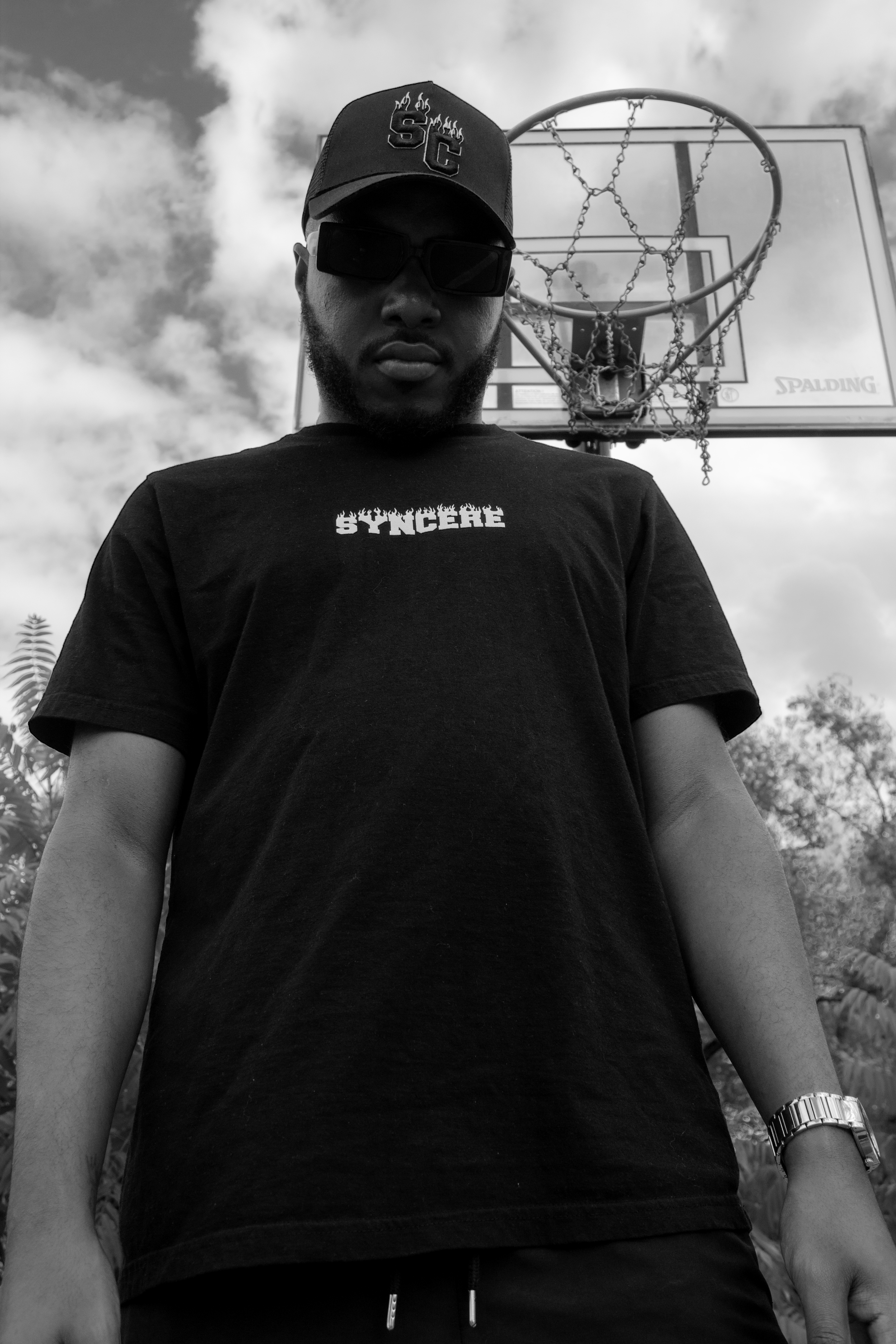 A man wearing a cap with flames and the number 23, sunglasses, a black t-shirt with the word 'SYNERGE' on it, and a watch, standing in front of a basketball hoop outdoors under cloudy skies.