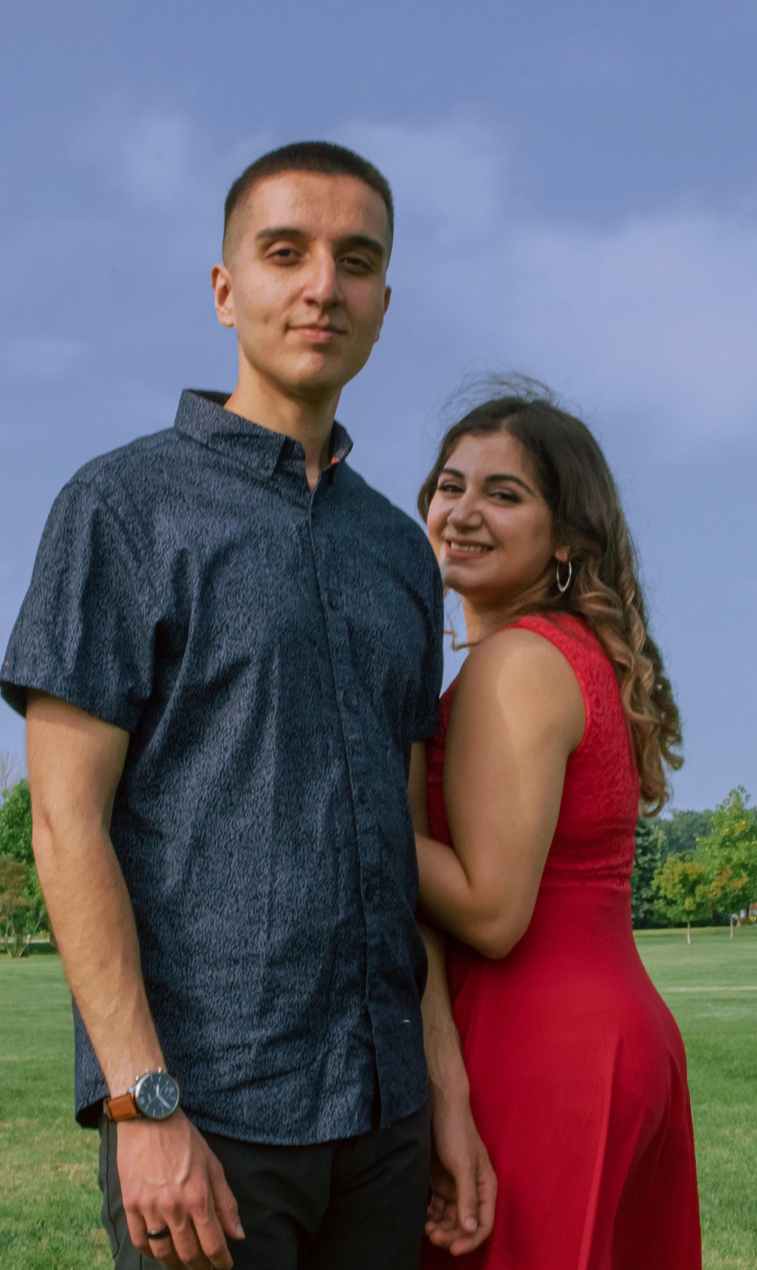 A young man and woman standing together outdoors under a cloudy sky, with trees and a grassy field in the background. The man is wearing a short-sleeved, button-up shirt and a watch, and the woman is wearing a sleeveless red dress.
