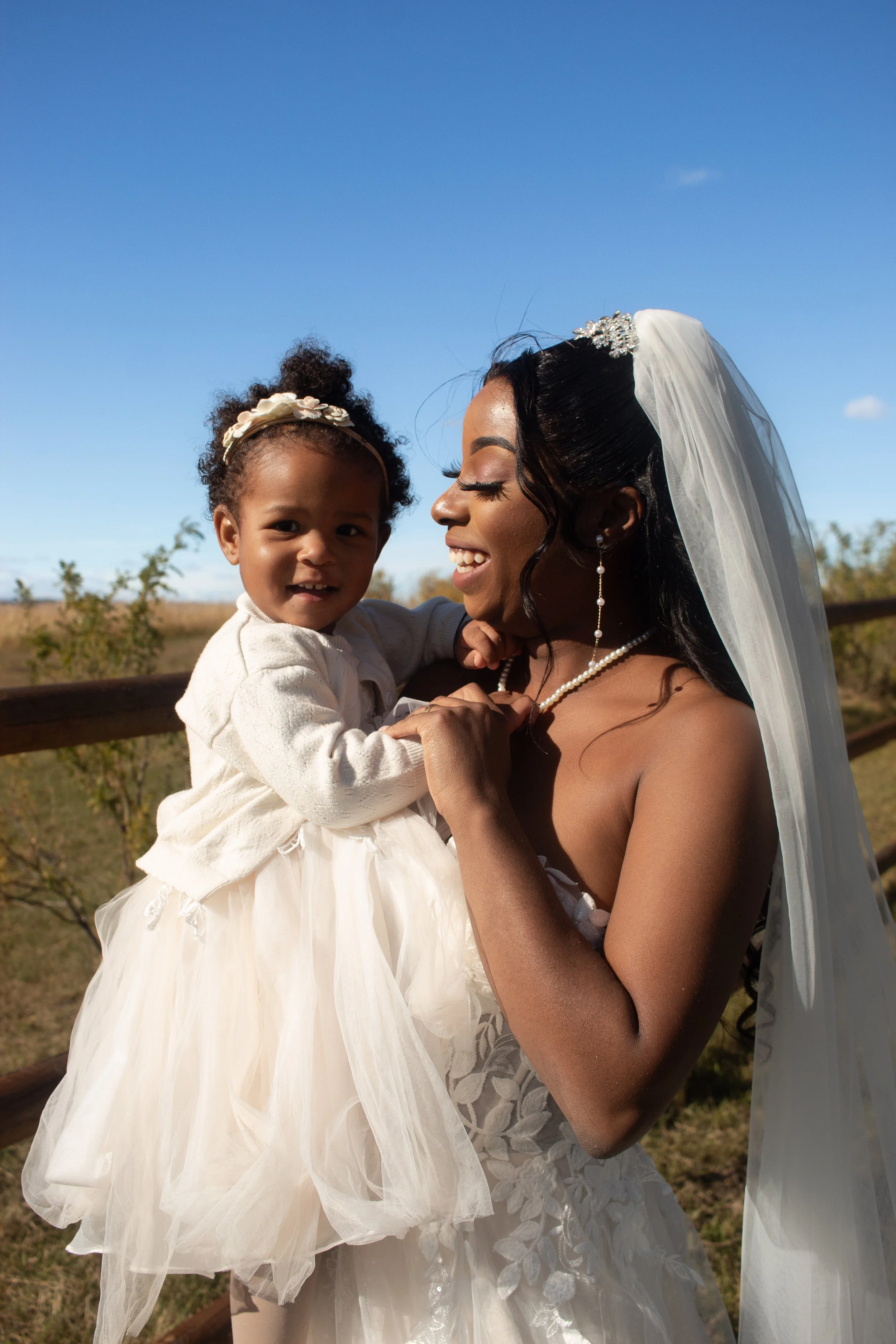 A woman in a wedding dress holding a smiling young girl in a white dress outdoors with a blue sky in the background.