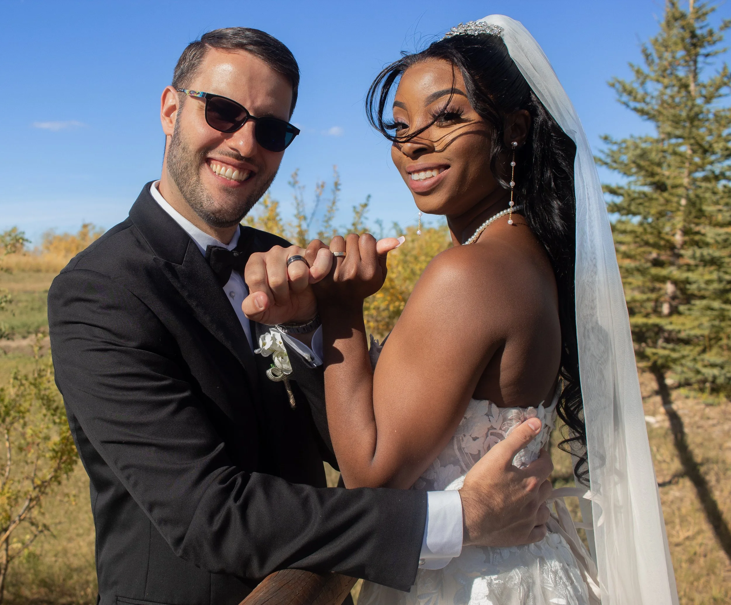 A smiling couple, a man in a black tuxedo with sunglasses and a woman in a wedding dress and veil, holding hands outdoors under a blue sky with trees in the background.