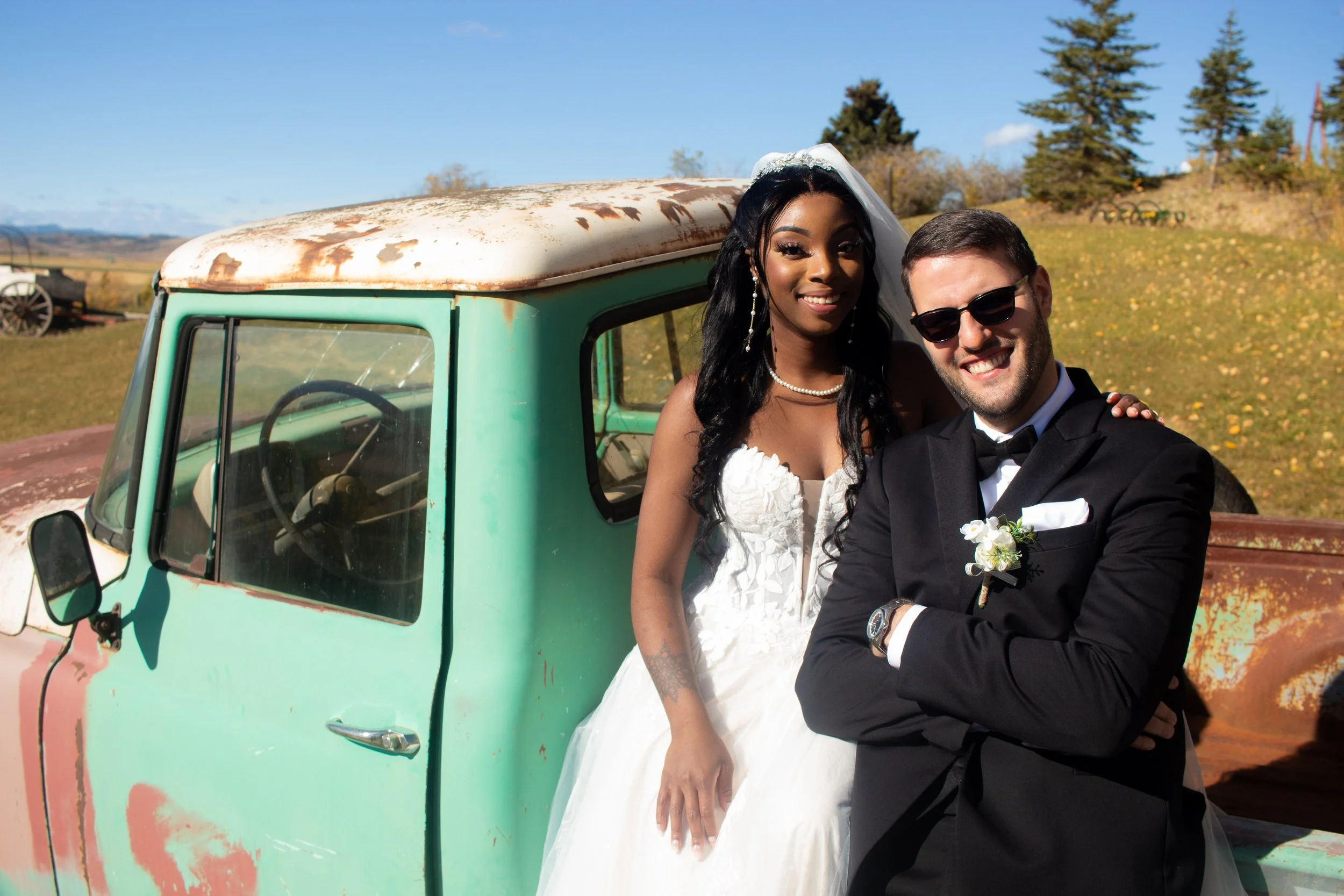 A bride and groom in wedding attire taking a selfie outdoors, standing beside a vintage, weathered green vehicle with a rusty roof, on a sunny day with clear blue skies.