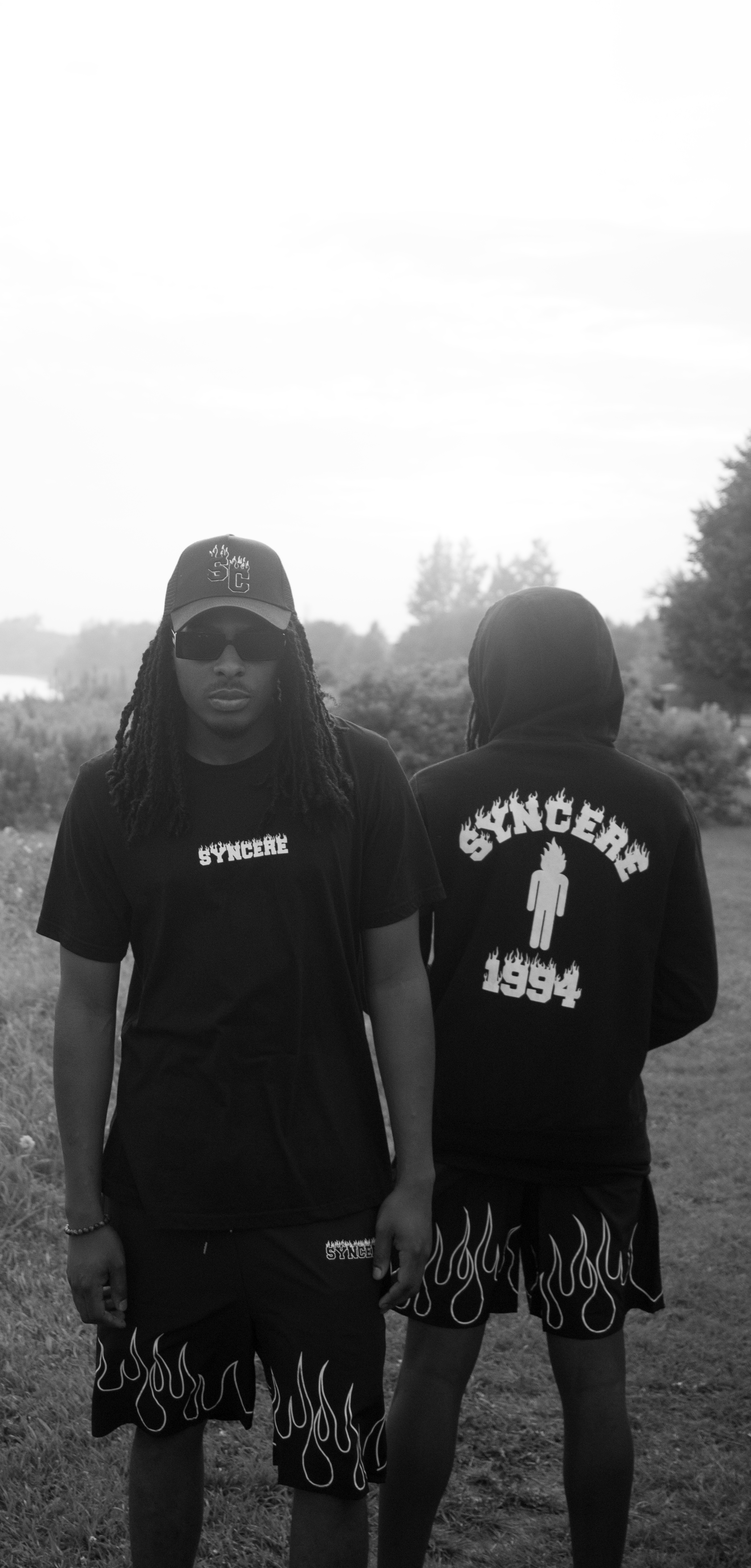 Two young men standing outdoors on a grassy area, wearing matching black T-shirts and shorts with flame patterns. One faces forward wearing sunglasses and a cap, the other has his back turned.