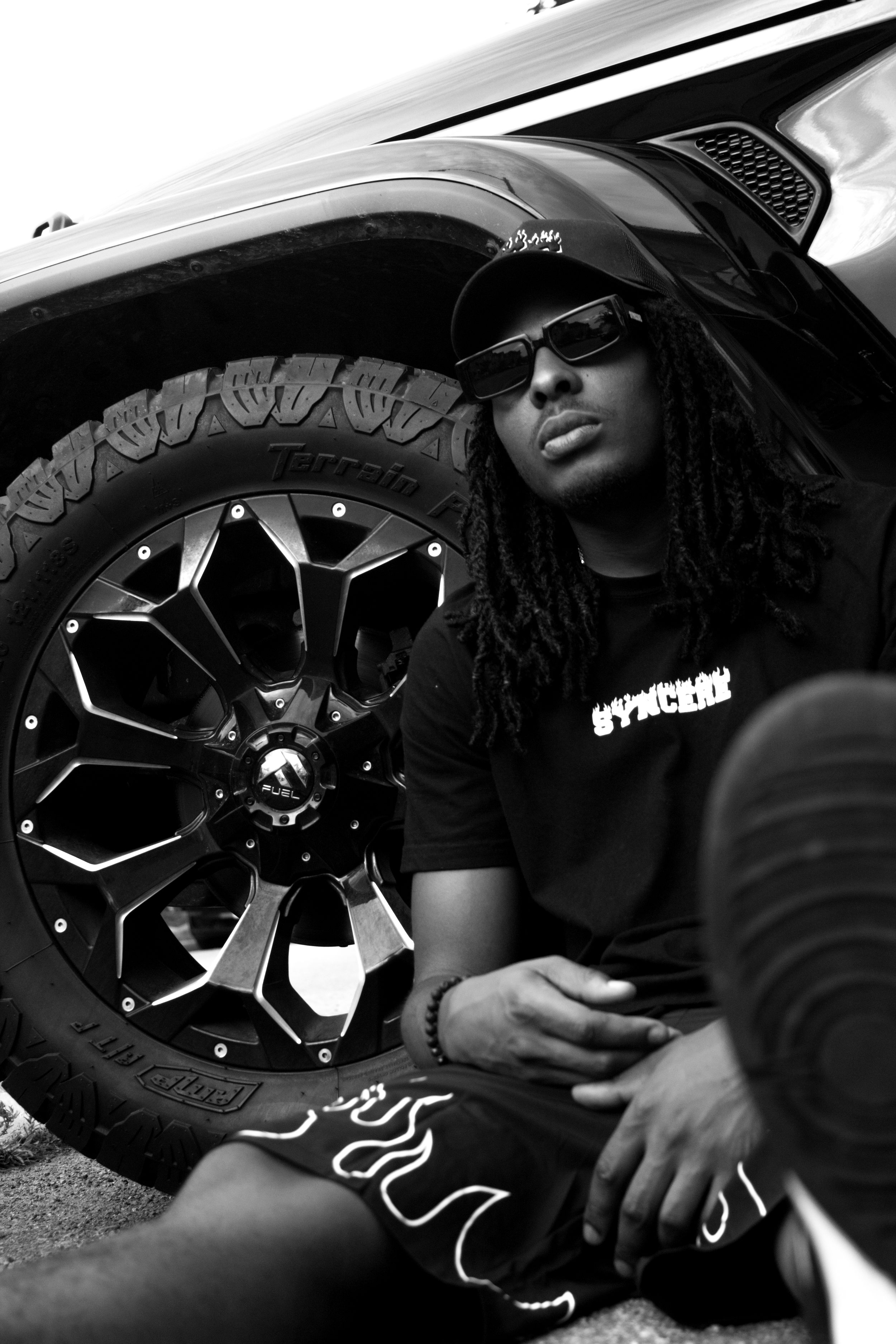 A person with dreadlocks wearing sunglasses and a cap, sitting beside a large off-road tire, with a part of a vehicle's side and wheel well visible in the background.