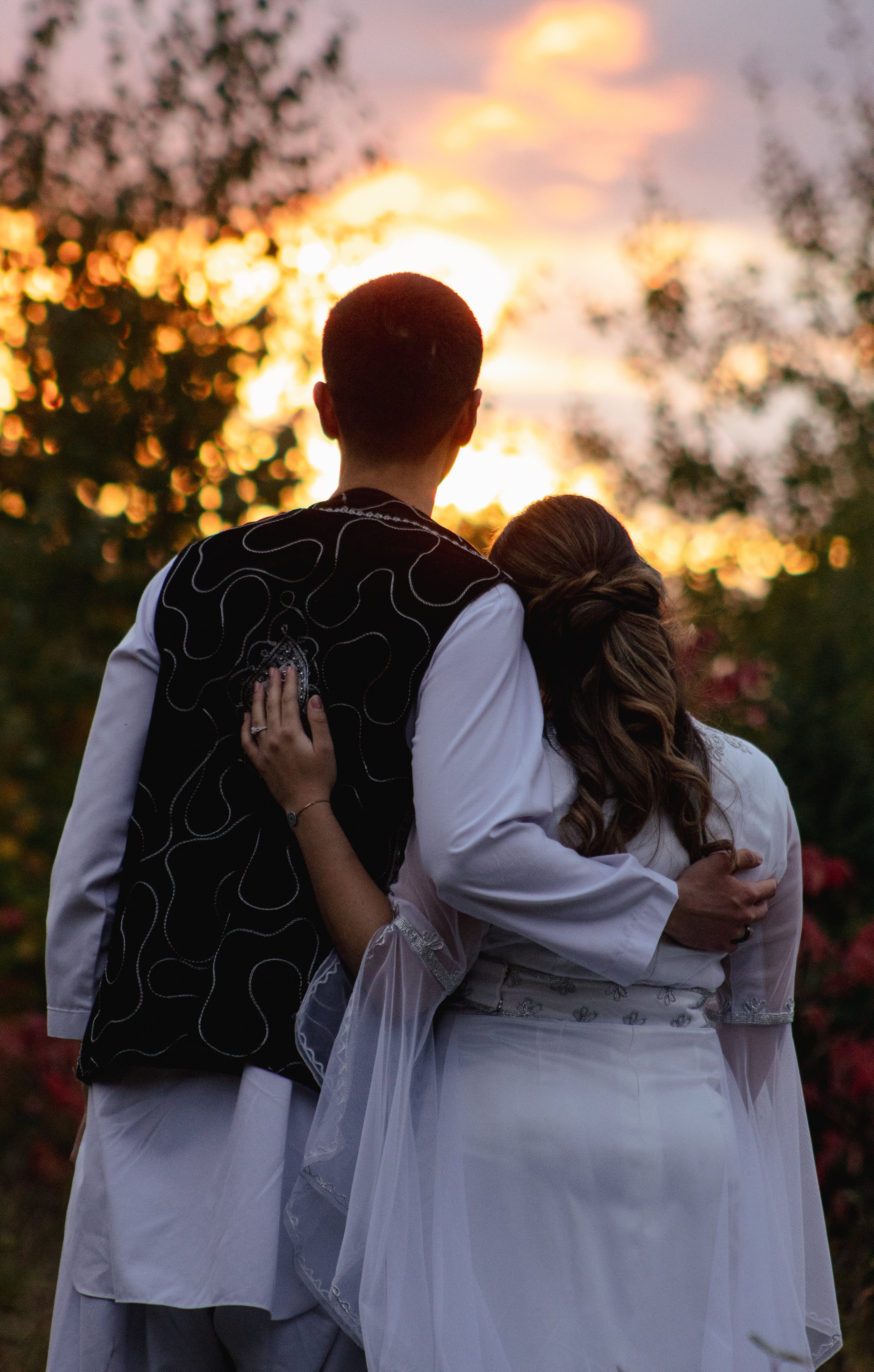 A couple with their arms around each other, standing outside during sunset, with trees and a colorful sky in the background.