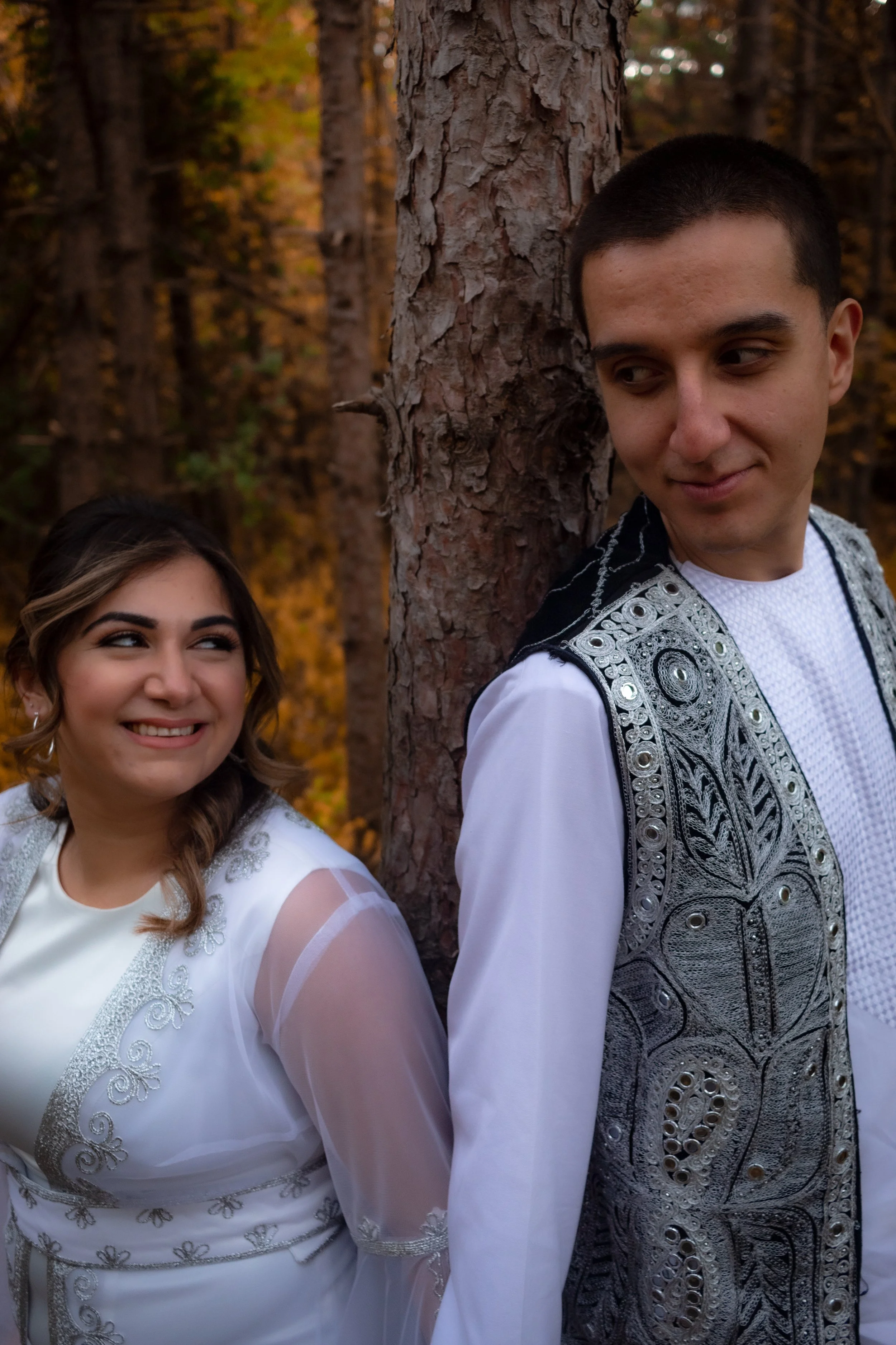 A young woman and a young man are standing outdoors in a forest with autumn foliage, leaning against a tree trunk, looking at each other and smiling. The woman has curly hair and is wearing a white dress with silver embroidery, and the man has short 